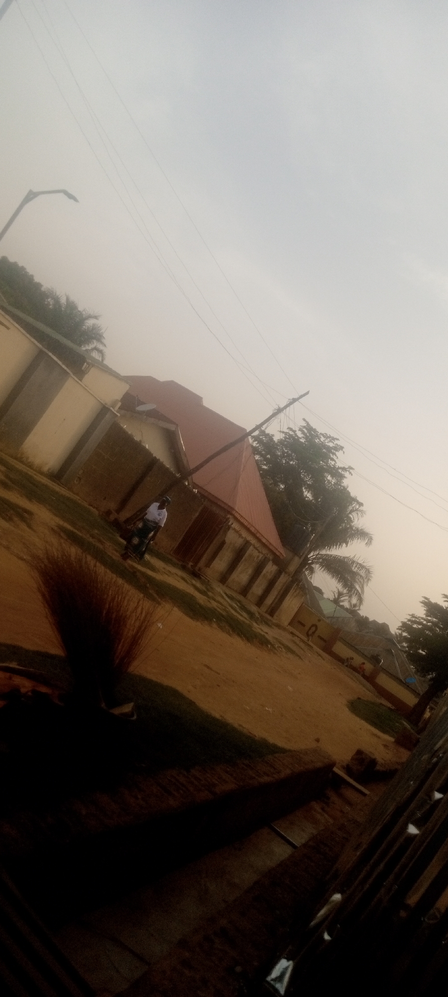 The image shows a residential street scene in what appears to be Narayi, Nigeria. The image is taken from a low angle, looking upwards and slightly to the side. The background features a building with a red roof, partially obscured by trees and a concrete wall. A man on a bicycle is visible in front of the building. An electrical pole with wires is overhead. A streetlight is visible in the upper left corner of the image. The sky is overcast. There are shadows and what appears to be a fence in the foreground. The ground is dirt or gravel. The overall lighting suggests either early morning or late afternoon.