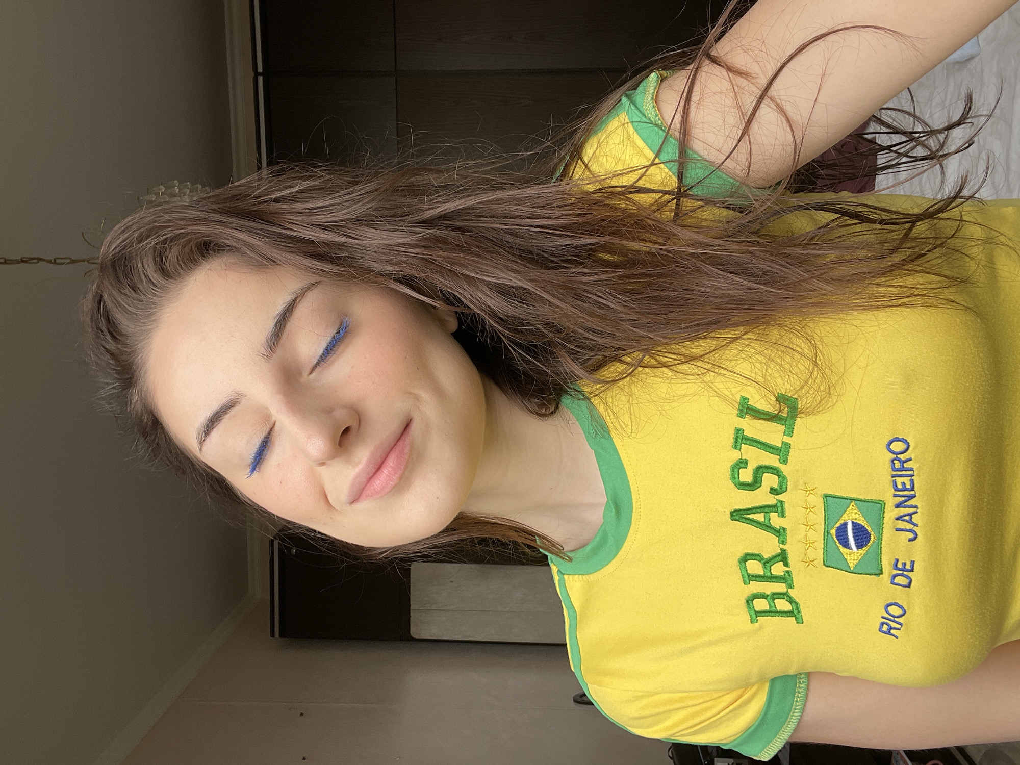 A young woman is captured in a close-up, chest-up indoor shot, lying down with her head tilted to her left. Her long, wavy brown hair is spread out around her. She has a serene expression, with her eyes gently closed and a soft smile gracing her lips, conveying a sense of peace and contentment.
She is wearing a vibrant yellow t-shirt with green trim along the neckline and sleeves. The t-shirt features prominent green embroidery, including the word "BRASIL" spelled vertically, four stars, a rectangular Brazilian flag emblem, and the words "RIO DE JANEIRO" also spelled vertically. A distinctive detail is the bold blue eyeliner visible on her closed eyelids. Her cheeks show a faint flush of blush.
The background elements are indistinct, suggesting an indoor setting with dark walls or furniture and some lighter-toned objects, including a metallic chain visible in the upper left. The photograph appears to be taken in natural or soft artificial light. The activity is one of repose, possibly resting or relaxing. Despite the Brazilian theme of her attire, the location of this media file is Moscow, Russia.