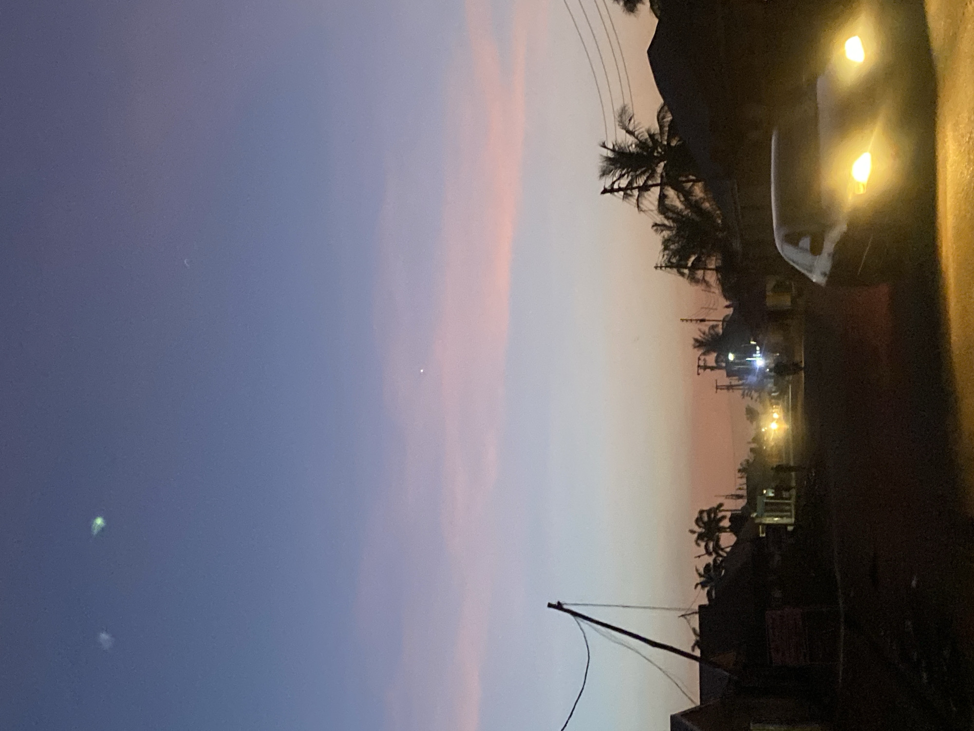 The image depicts a twilight scene in Sabon Tasha, Nigeria. The sky transitions from a deep blue-purple to light pink-orange on the horizon. Palm trees are silhouetted against the fading light. Power lines stretch across the sky. Buildings are visible on the right side of the image. A vehicle with illuminated headlights is parked on the right. Streetlights dot the road.