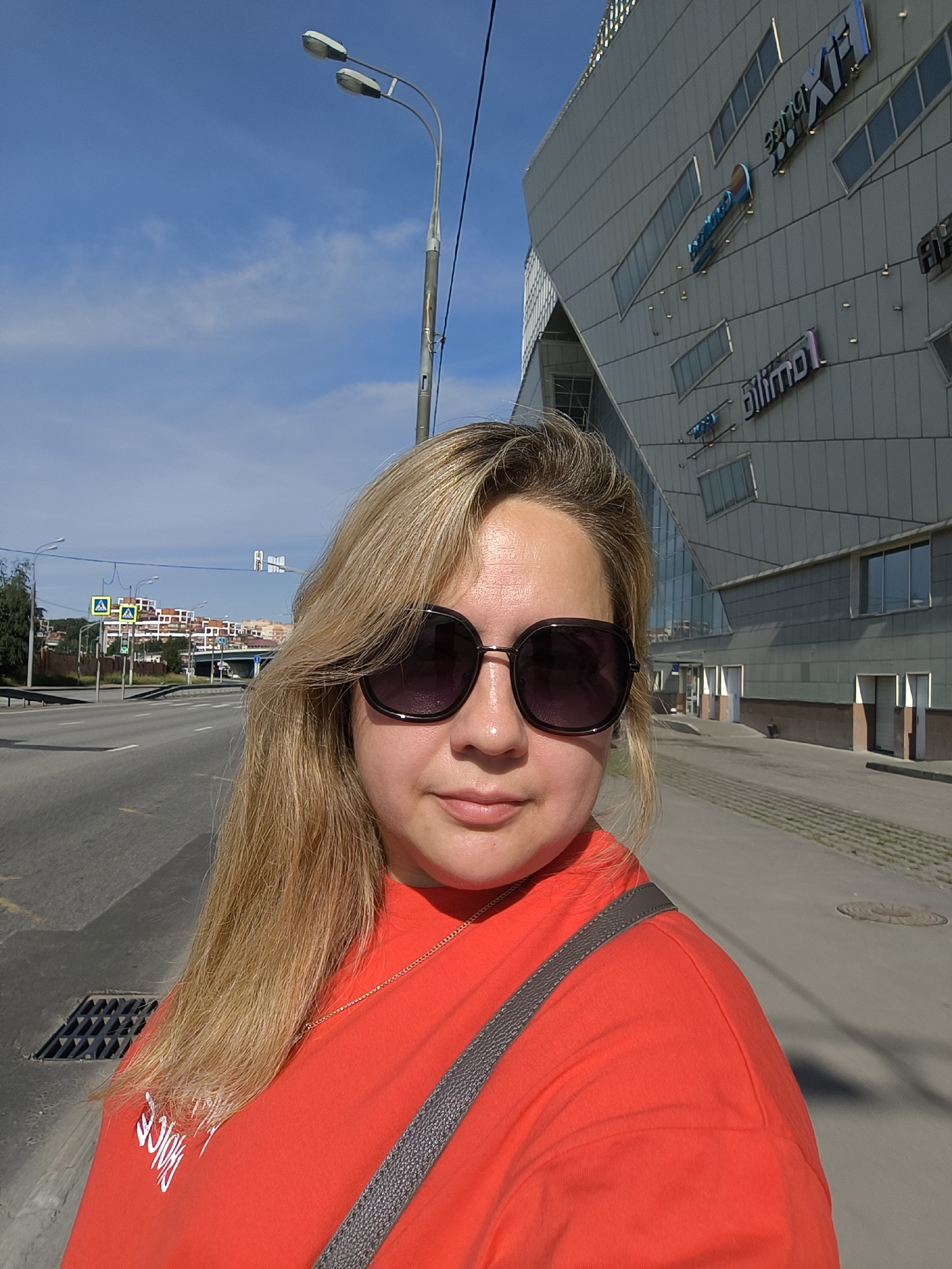 A woman with blonde hair, wearing sunglasses and a red shirt, stands in front of a modern building. The building has a distinctive angular design with several windows and visible signage, including blue lettering on the facade. A street runs parallel to the building, with a sidewalk in between. There are streetlights and traffic signals visible in the background. The sky is blue with a few wispy clouds. The scene appears to be an urban environment on a sunny day.