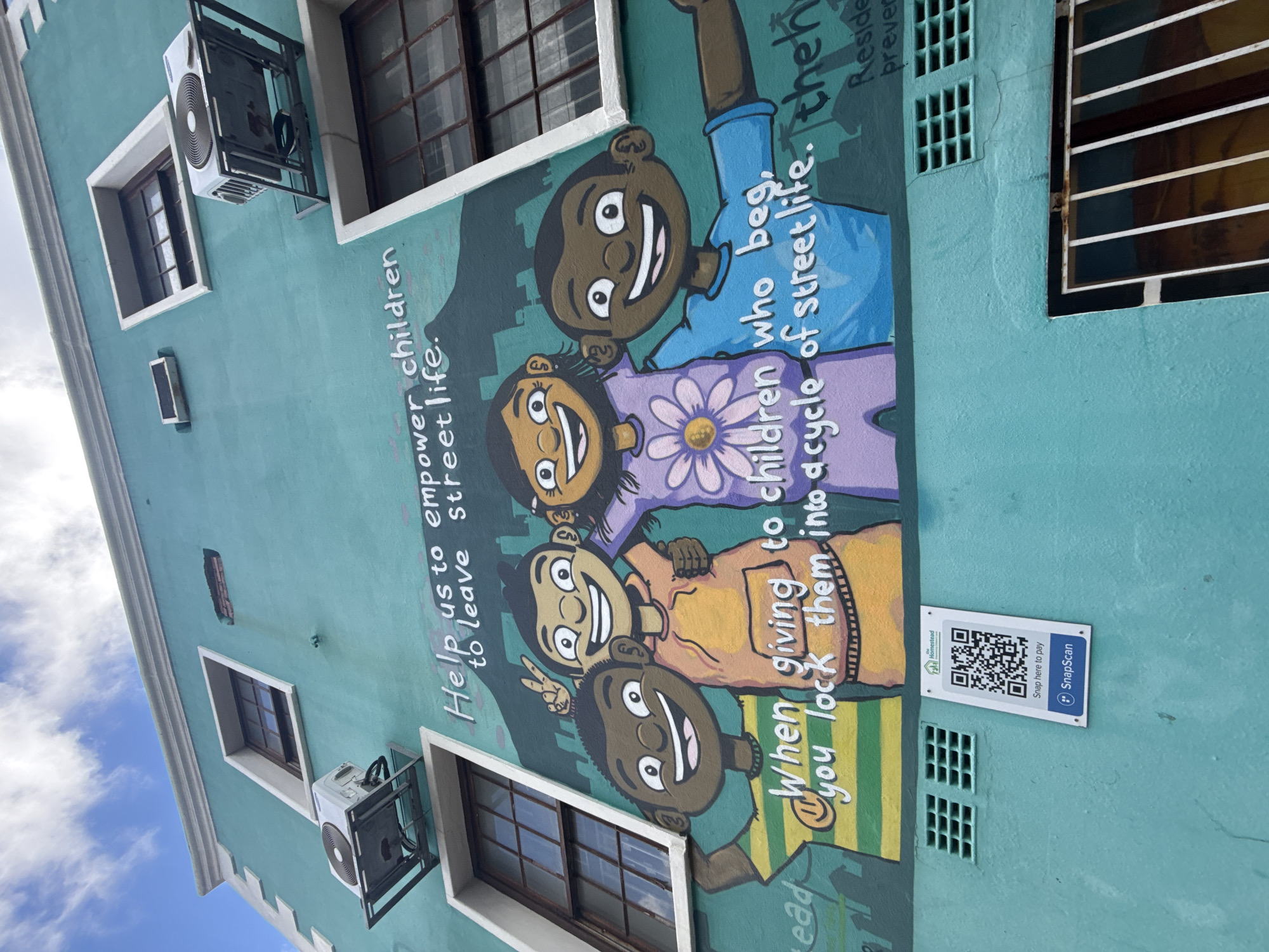 The image shows a brightly colored mural on the side of a building in what appears to be the city of Cape Town, South Africa. The mural depicts cartoon-like images of children with smiling faces and outstretched hands, along with the text: "Help us to empower children to leave street life," and "When giving to children who beg, you lock them into a cycle of street life." Adjacent to the mural is a window and an air conditioning unit. Also visible is a sign with a QR code and the text "Snap here to pay" and "SnapScan." The building is painted a light turquoise color, and the sky is visible in the background.