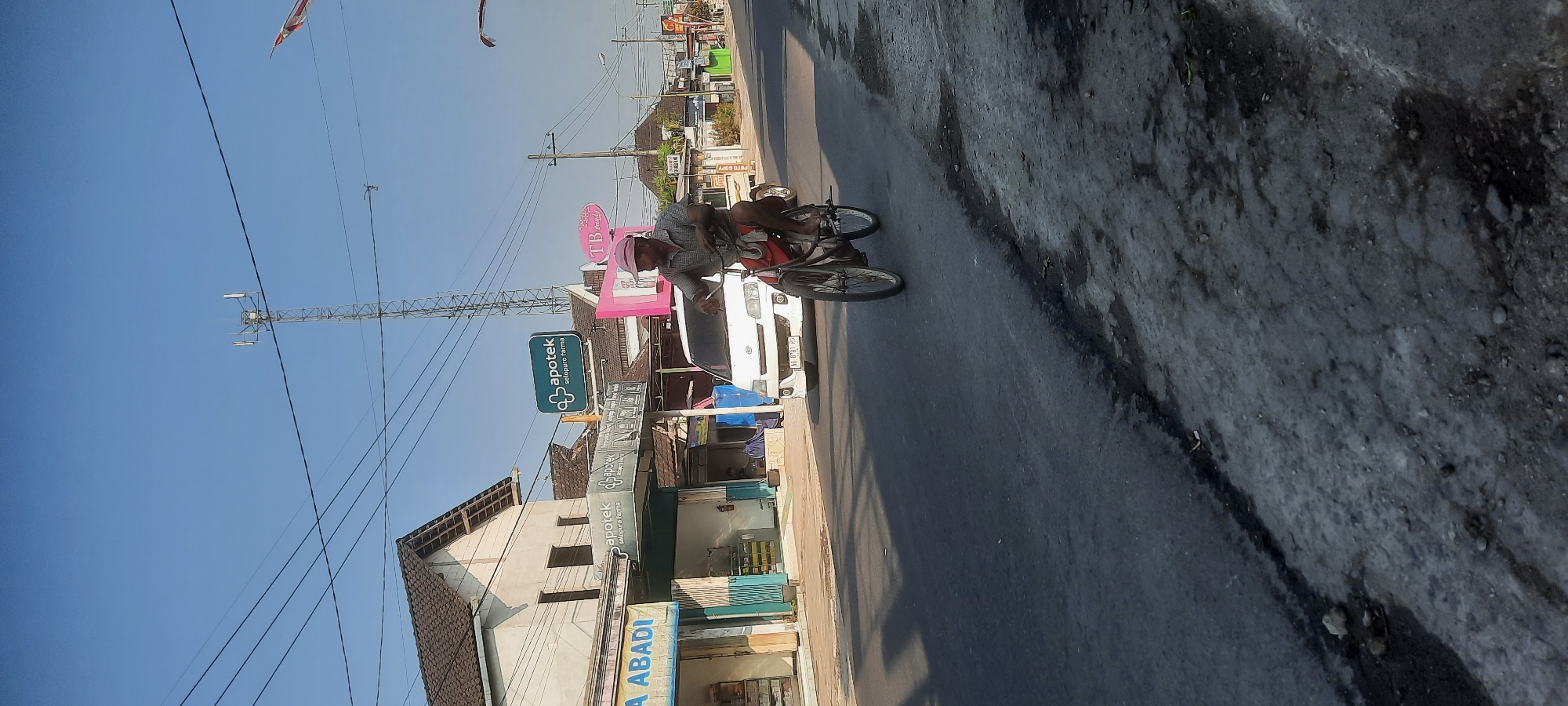 In Selopuro, Indonesia, a man is riding a bicycle on a paved road. The man is wearing a pink cap, a plaid shirt, and red shorts. He is on a bicycle with two wheels, and the front wheel appears to be larger than the back wheel. He is riding on the right side of the road, which is cracked and appears to be in disrepair in some areas. Behind him, a white van is also on the road, driving in the same direction. On the left side of the road are buildings, including a building with a sign that reads "ApoteK setopuro farma". There are also power lines visible across the top of the image. The sky is clear and blue. A telecommunications tower is visible in the background.