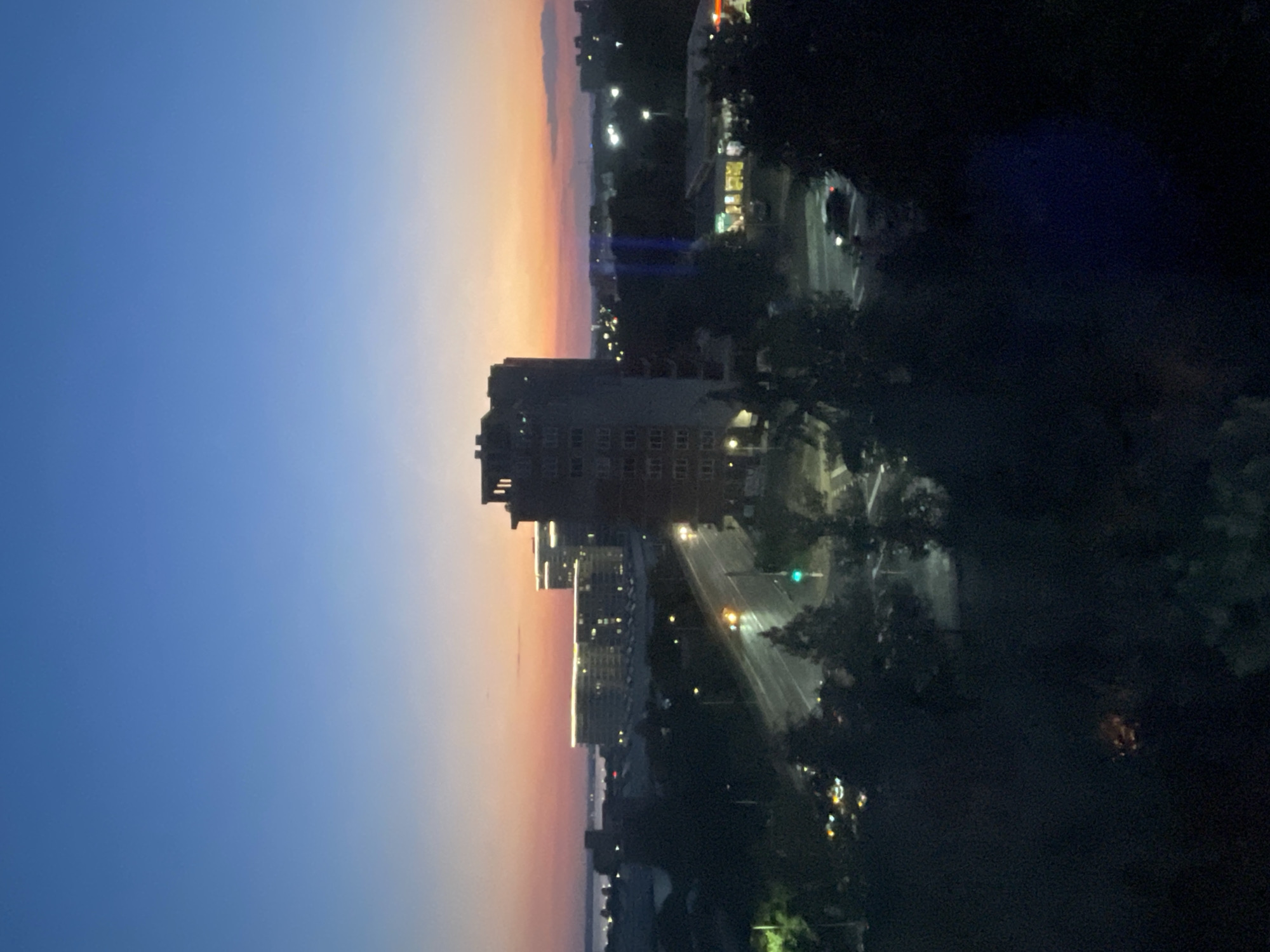 This image depicts a twilight scene in Burgas, Bulgaria, likely from an elevated vantage point. The sky transitions from a deep blue at the top to hues of orange and pink near the horizon, indicating either dawn or dusk. In the foreground, dark silhouettes of trees partially obscure the view.
Dominating the mid-ground is a multi-story building with numerous windows. To its left, a road is visible, illuminated by streetlights and the headlights of a few vehicles. A traffic light shows a green signal. Further in the distance, several modern buildings with illuminated windows are clustered, some appearing to be a complex of hotels or apartments, and a body of water, likely the Black Sea, is visible on the far left. The overall atmosphere is calm, with the city lights starting to twinkle as daylight fades. There are no visible people in the image.