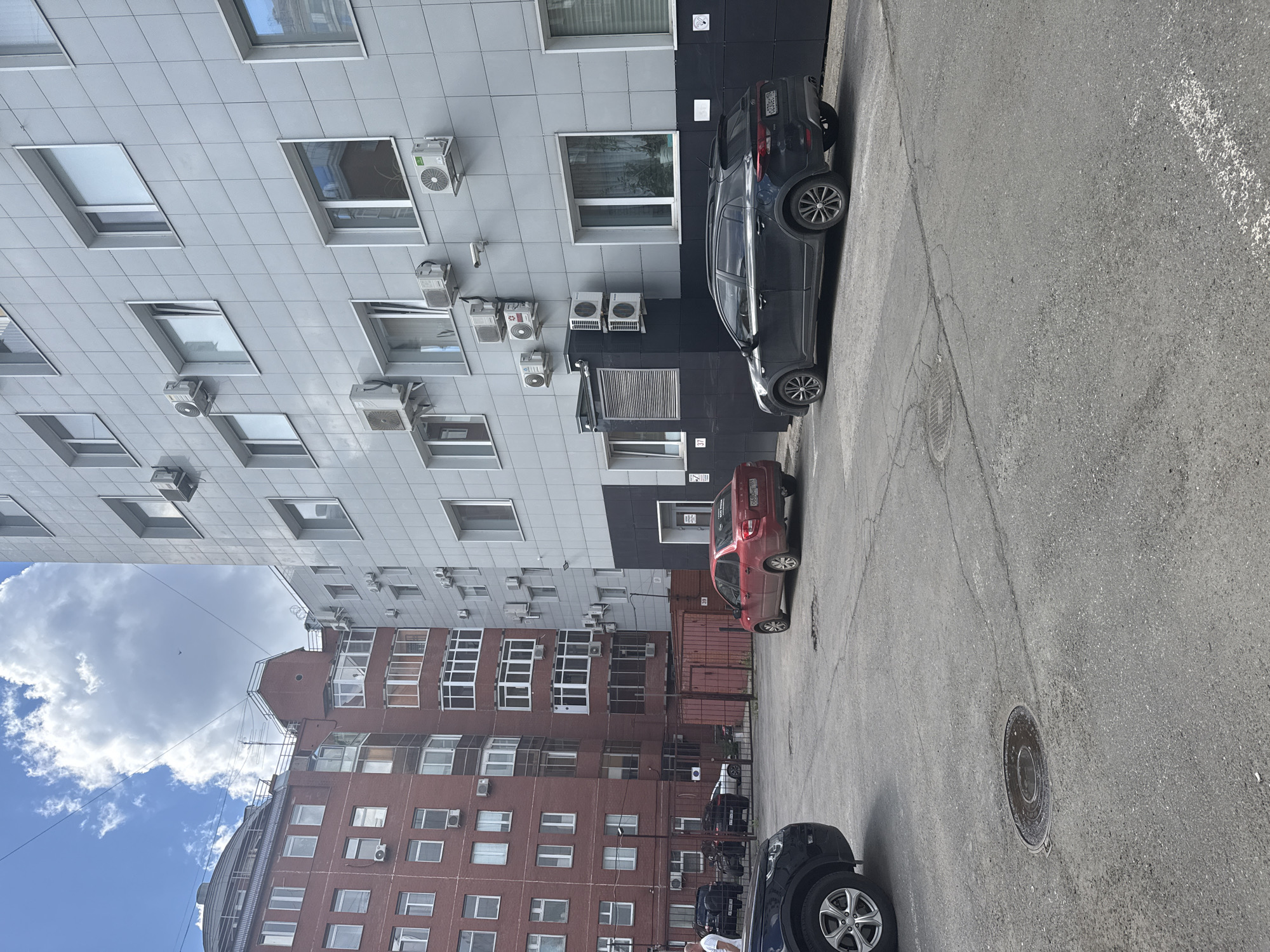 The image shows an outdoor scene in Perm, Russia, likely a parking lot or courtyard adjacent to buildings. In the foreground, a gray asphalt surface with visible cracks and a manhole cover is present. A black SUV is parked on the right side, facing away from the camera. To the left and slightly further back, a red hatchback is also parked. Behind the cars, a dark fence encloses an area with some red industrial containers. In the background, two multi-story buildings stand side-by-side. The building on the left is made of red brick and features numerous windows and several visible air conditioning units. The building on the right is clad in gray tiles and also has many windows and air conditioning units. The sky above is blue with large white clouds, and thin wires stretch across the frame. The overall impression is an urban setting on a daytime.