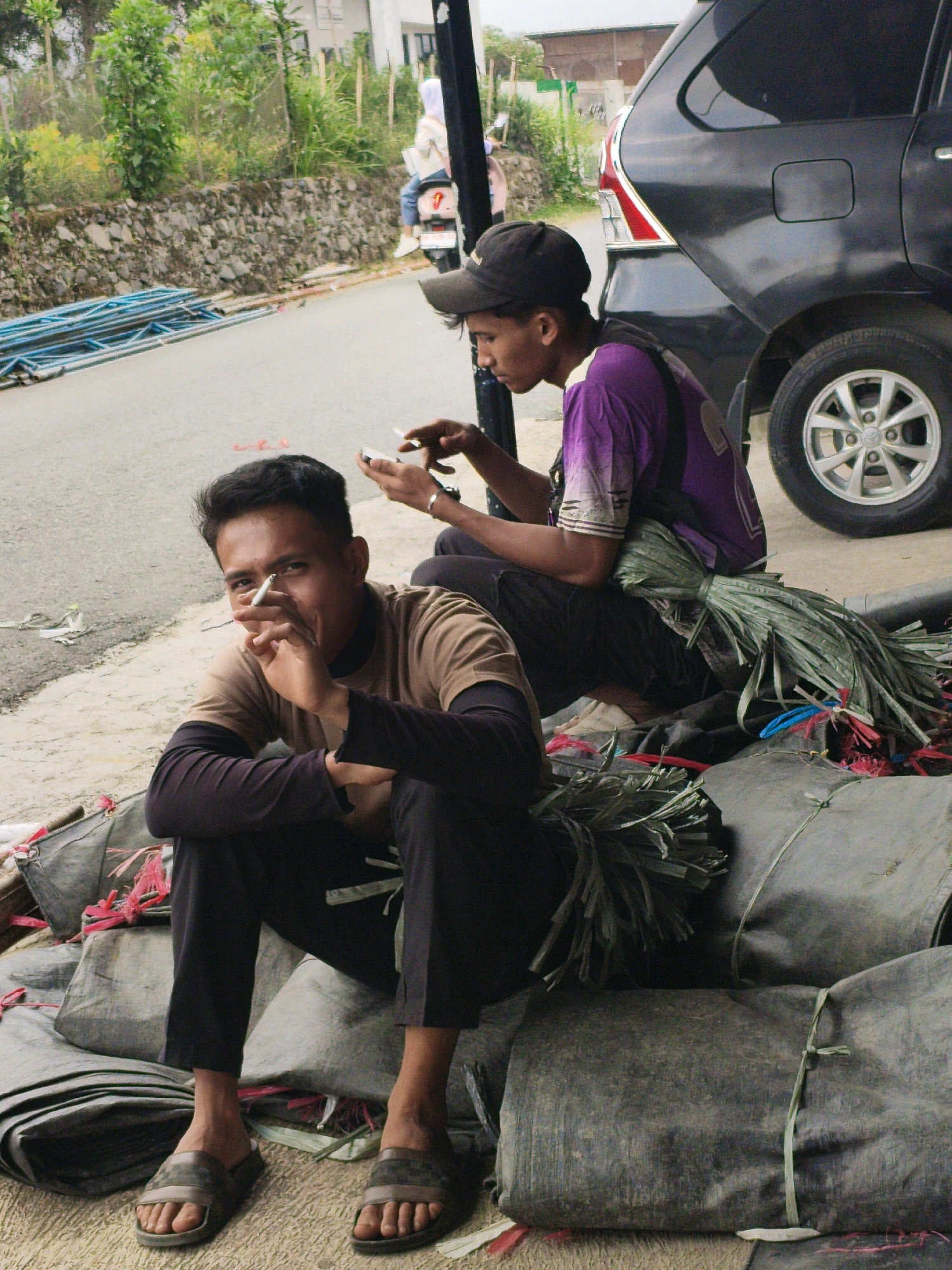 The image depicts two young men sitting on large, bundled sacks of what appear to be dried grass or reeds, located on the side of a paved road in Lindung Jaya, Indonesia. In the foreground, a man in a tan long-sleeved shirt and black pants is smoking a cigarette and looking towards the camera. He is wearing dark sandals. Behind him and to his right, another young man wearing a purple shirt and a black cap is looking down at his phone. A dark-colored car is parked to the right of the frame. In the background, there is a stone wall, a building, and some greenery, suggesting an outdoor, semi-urban or rural setting. The overall atmosphere appears casual and somewhat unkempt, with the bundled materials and scattered debris on the roadside.