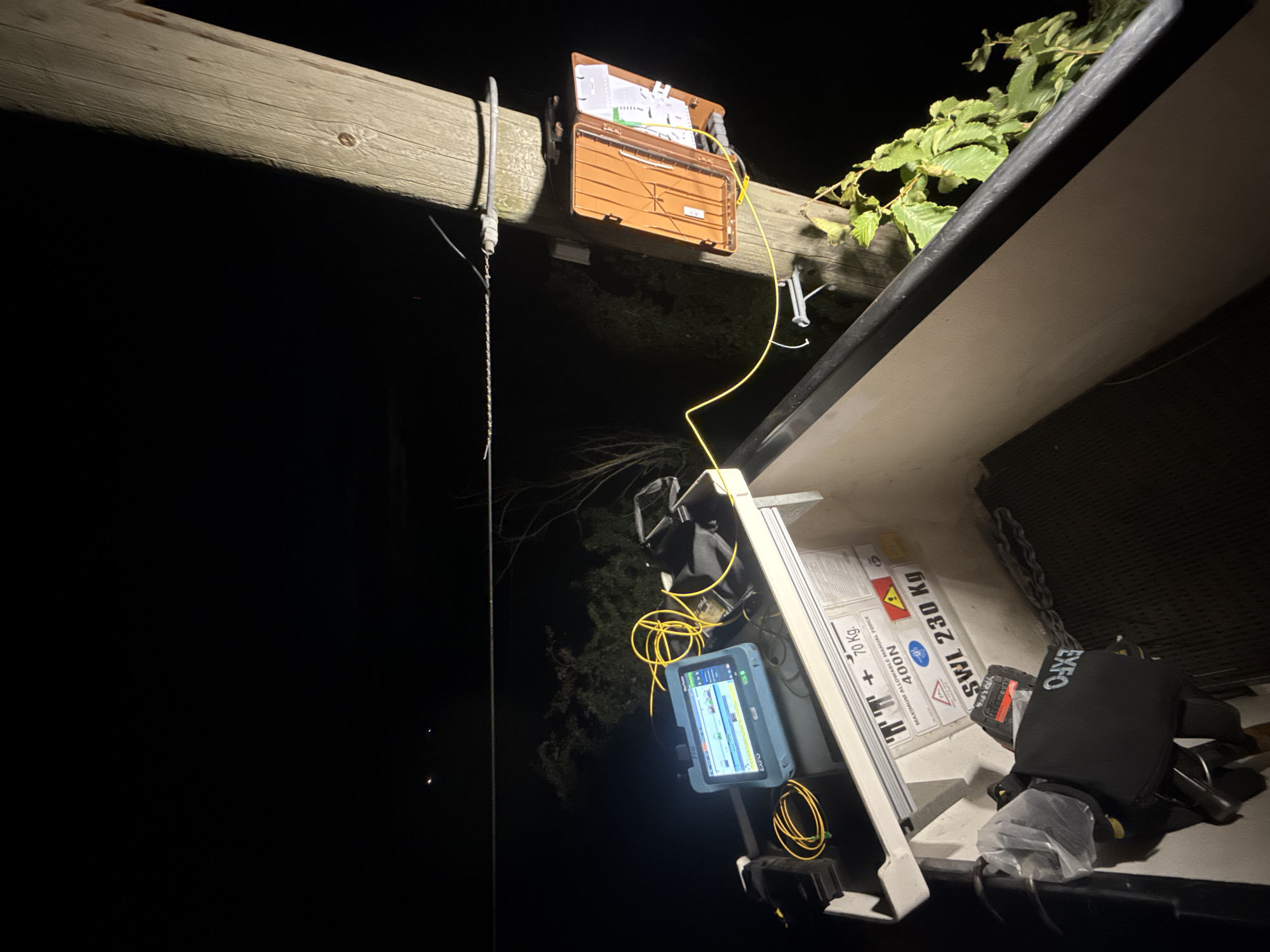 The image depicts an outdoor scene at night, possibly in the city of East Suffolk, United Kingdom, where fiber optic cable work is in progress. A wooden utility pole is prominently featured, with an orange fiber optic distribution box mounted on it. Yellow fiber optic cables are visible, connecting the distribution box to testing equipment. A blue fiber optic tester, identified as an EXFO MaxTester, is set up on a white platform or bucket. Beside the tester is a black carrying case with the EXFO logo. Warnings and safety labels are affixed to the platform, indicating a maximum load of 70 kg, a working load limit of 230 kg, and a maximum allowable manual force of 400N, along with hazard symbols. The surrounding environment is dark, with some foliage visible, and the sky suggests a clear night. The overall scene conveys a sense of active infrastructure maintenance being conducted after dark.