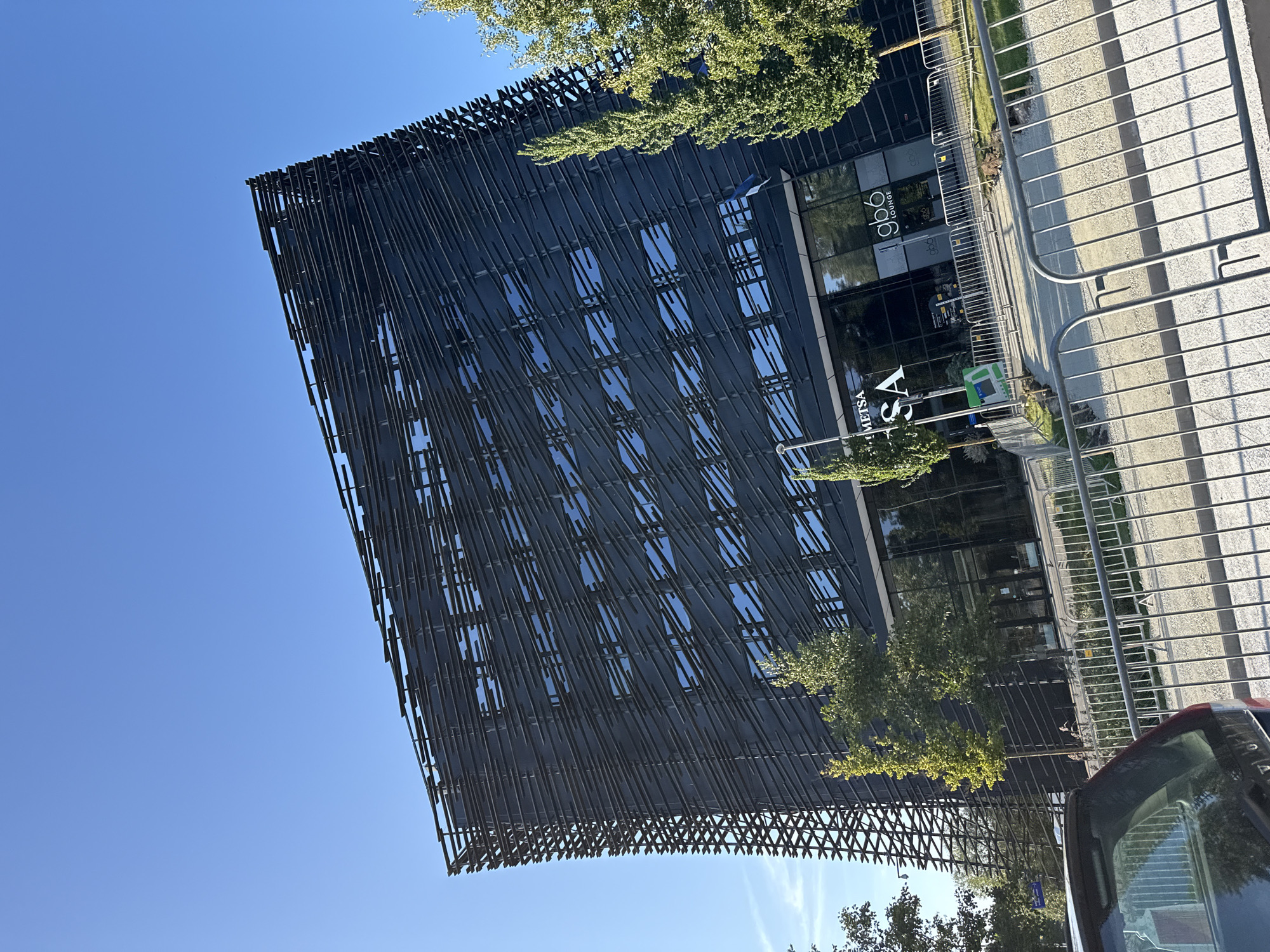 The image shows a modern, angular building with a dark, slatted facade, likely made of metal. The building appears to be a commercial or office space, with large glass doors and windows at street level. Signs on the building indicate the presence of "METSA" and "gb6 LOUNGE". The building is surrounded by trees, some of which are green and leafy, suggesting it is daytime and possibly spring or summer. A silver car is partially visible in the foreground, parked behind a metal fence. The sky is clear and blue. The architectural style of the building and the presence of the Estonian flag (a white cross on a blue background) visible on a flagpole near the entrance strongly suggest the location is Tallinn, Estonia. Specifically, this building is the Metsä Wood headquarters, located in the Lasnamäe district of Tallinn.