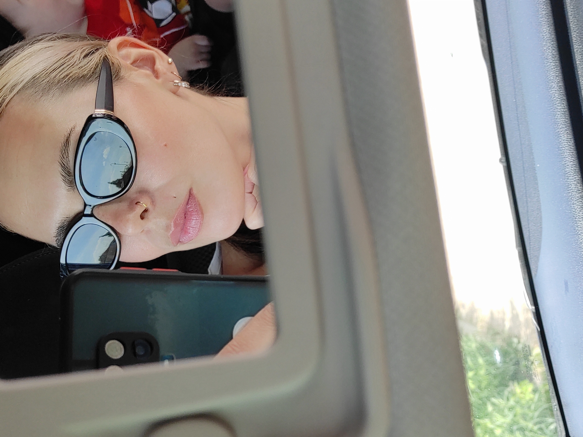 A selfie of a light-skinned person with blonde hair, wearing sunglasses and lip gloss, reflected in a vehicle's side mirror. The subject has a nose ring and earrings. The person is holding a smartphone with a black case. The view outside the vehicle window shows greenery. No specific details indicate the location is Chaikovsky, Russia.