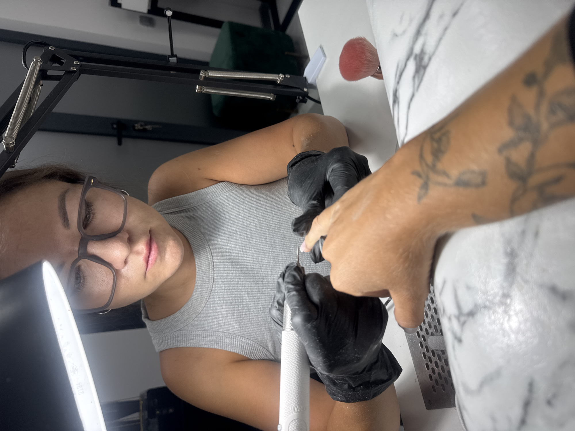 A woman, wearing black gloves and a grey tank top, is using an electric nail file to work on a client's fingernail. The woman has glasses and is focused on the task. The client's hand with a tattoo is resting on a white surface with a marble pattern. A pink makeup brush is visible on the table. The scene appears to be a nail salon. The location is Budva, Montenegro.