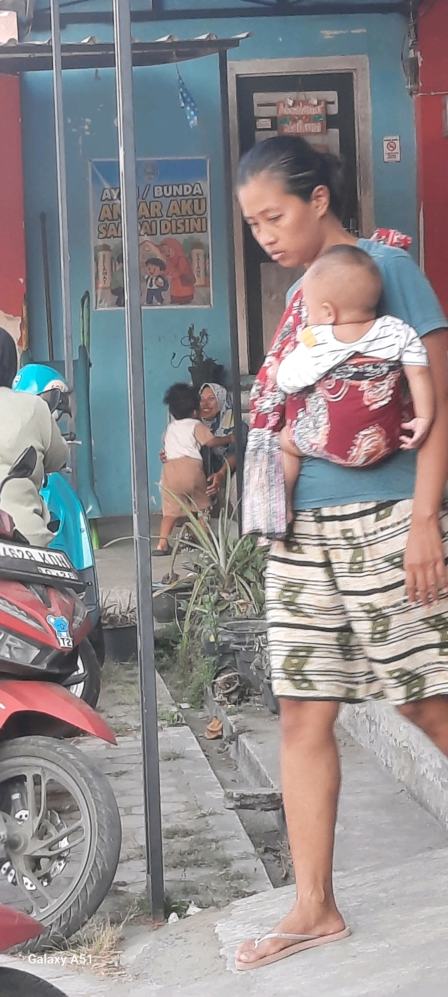 In Selopuro, Indonesia, a woman wearing a turquoise t-shirt and patterned shorts carries a baby wrapped in a red and white patterned cloth on her back. To the left, a red motorcycle is parked near a turquoise scooter. Behind the woman, a smaller child in a light brown outfit is being hugged by another woman wearing a headscarf. A blue wall with a poster featuring animated characters serves as the backdrop, and a metal pole stands prominently in the foreground. The scene appears to be on a street or pathway with potted plants and concrete steps.