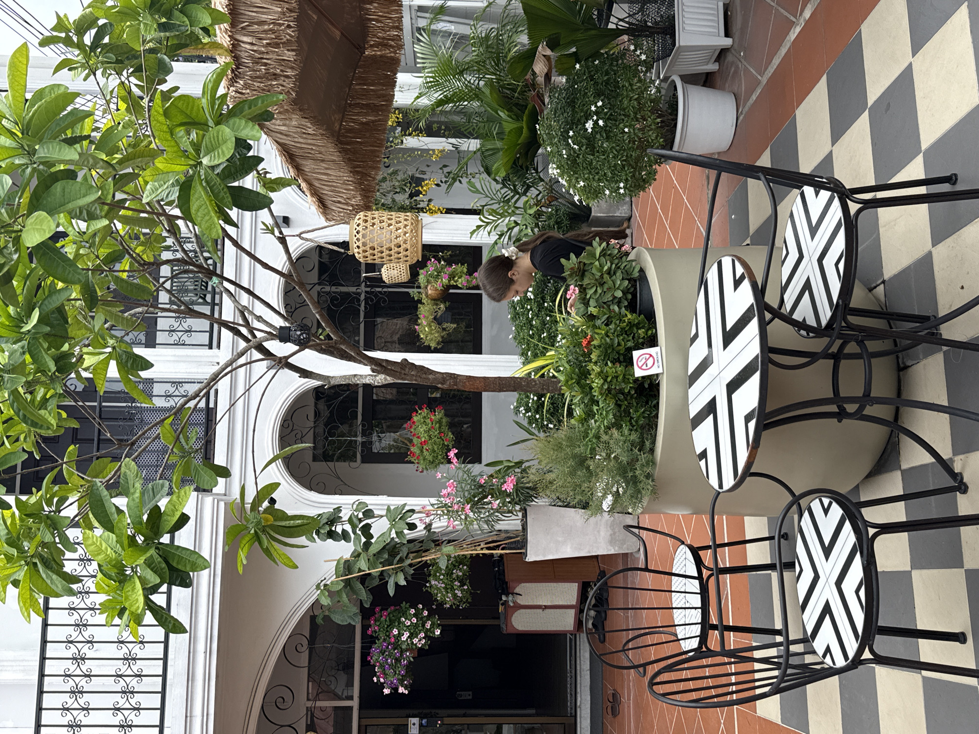 The image captures an outdoor courtyard area, possibly a cafe or restaurant, in Bangkok, Thailand. A young woman with her hair tied back in a ponytail, wearing a dark t-shirt, is tending to plants in a circular planter. In the foreground, a black metal table with a patterned top and two matching chairs with patterned seats are arranged on a checkered tiled floor. The courtyard features lush greenery, with various potted plants and hanging baskets of flowers. A straw-covered umbrella provides shade. In the background, there are arched doorways and windows with decorative railings, indicative of a traditional architectural style. A "No Smoking" sign is visible on the planter. The overall atmosphere is serene and inviting, with a focus on natural elements and outdoor seating.