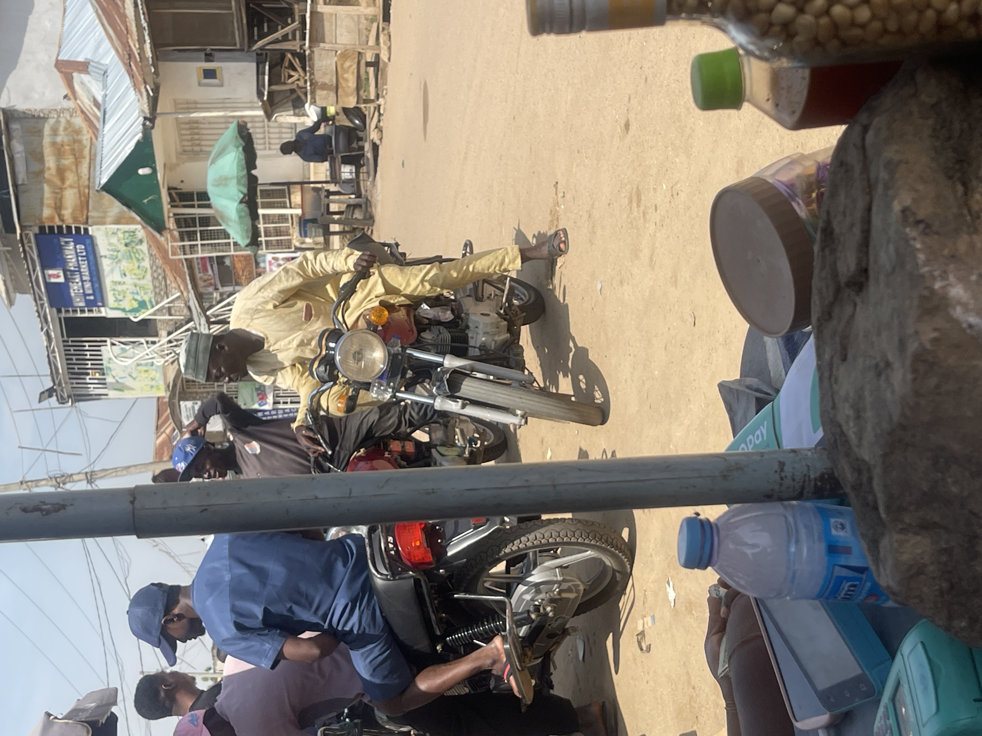 The image depicts a street scene in Abuja, Nigeria, likely a market or commercial area, under a bright, sunny sky. In the foreground, several objects are placed on a surface, including a plastic water bottle, a clear jar with a brown lid, a small brown bottle with a green cap, and a glass bottle containing what appears to be nuts or seeds. Also visible are parts of electronic devices, possibly payment terminals or mobile phones, and a large rock.
The middle ground is dominated by several people on motorcycles. One man, wearing a light yellow traditional outfit and a cap, is seated on a red motorcycle, holding the handlebars. To his left, another man in a blue shirt and cap is also on a motorcycle, appearing to be interacting with the man in yellow. Other individuals are visible on motorcycles in the background. The motorcycles are parked or moving on a dirt or paved road, typical of many streets in Nigerian cities.
The background shows a row of buildings with varying architectural styles. Some have corrugated metal roofs, while others have more permanent structures with windows and signs. One building has a prominent sign that reads "WHITEHEART PHARMACY & MINI-MARKET LTD." There are also awnings and stalls, suggesting commercial activity. The overall impression is a bustling, informal urban environment characteristic of parts of Abuja.