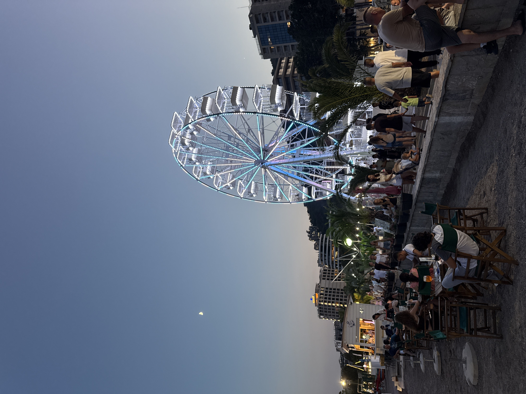 The image depicts a lively evening scene in Budva, Montenegro, with a large Ferris wheel illuminated by colorful lights dominating the sky. A crescent moon is visible in the dusky sky, suggesting it's either late afternoon or early evening. The Ferris wheel, a prominent attraction, features numerous gondolas and is adorned with a vibrant display of white, blue, and purple lights.
Below the Ferris wheel, a bustling promenade is filled with people enjoying the atmosphere. Many are seated at tables outside various establishments, appearing to be dining or socializing. Some individuals are standing and observing the surroundings. The area is lined with palm trees and modern buildings, indicating a developed urban waterfront. The ground is paved, with some areas appearing to be gravel.
The overall mood is festive and relaxed, with a sense of community gathering. The warm evening light and the illuminated Ferris wheel contribute to a pleasant ambiance. The image captures a moment of leisure and enjoyment in a popular tourist destination. No discernible text is visible in the image.