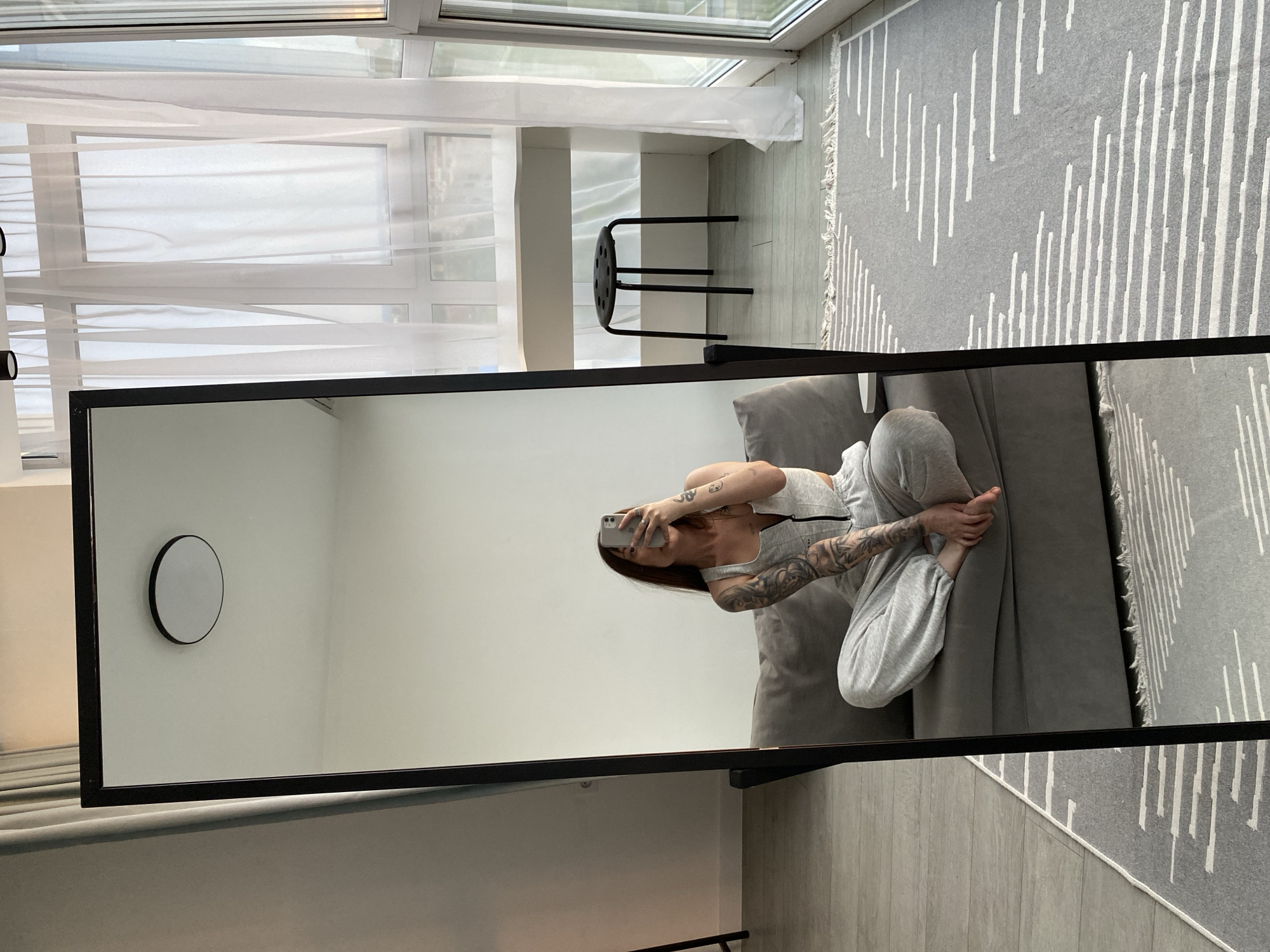 This media file is a photograph of a woman taking a mirror selfie in a well-lit room. The room appears to be a living area or bedroom with large windows covered by sheer white curtains, suggesting daytime with bright, possibly overcast, weather. A grey rug with a white abstract pattern covers a portion of the light-colored floor. In the background, a black stool with a circular seat and multiple legs is visible near the windows.
The woman, positioned on a grey sofa, is wearing a light grey athletic top and matching sweatpants, accentuating a casual and relaxed mood. She has visible tattoos on her arms. She is holding a smartphone in her right hand, angled to capture her reflection in a large, rectangular mirror with a black frame that dominates the foreground of the image. The mirror reflects the woman, the sofa, and a portion of the room, including a small circular wall-mounted mirror. The overall impression is one of a candid, personal moment captured in a modern, minimalist living space. There is no visible text in the image.
