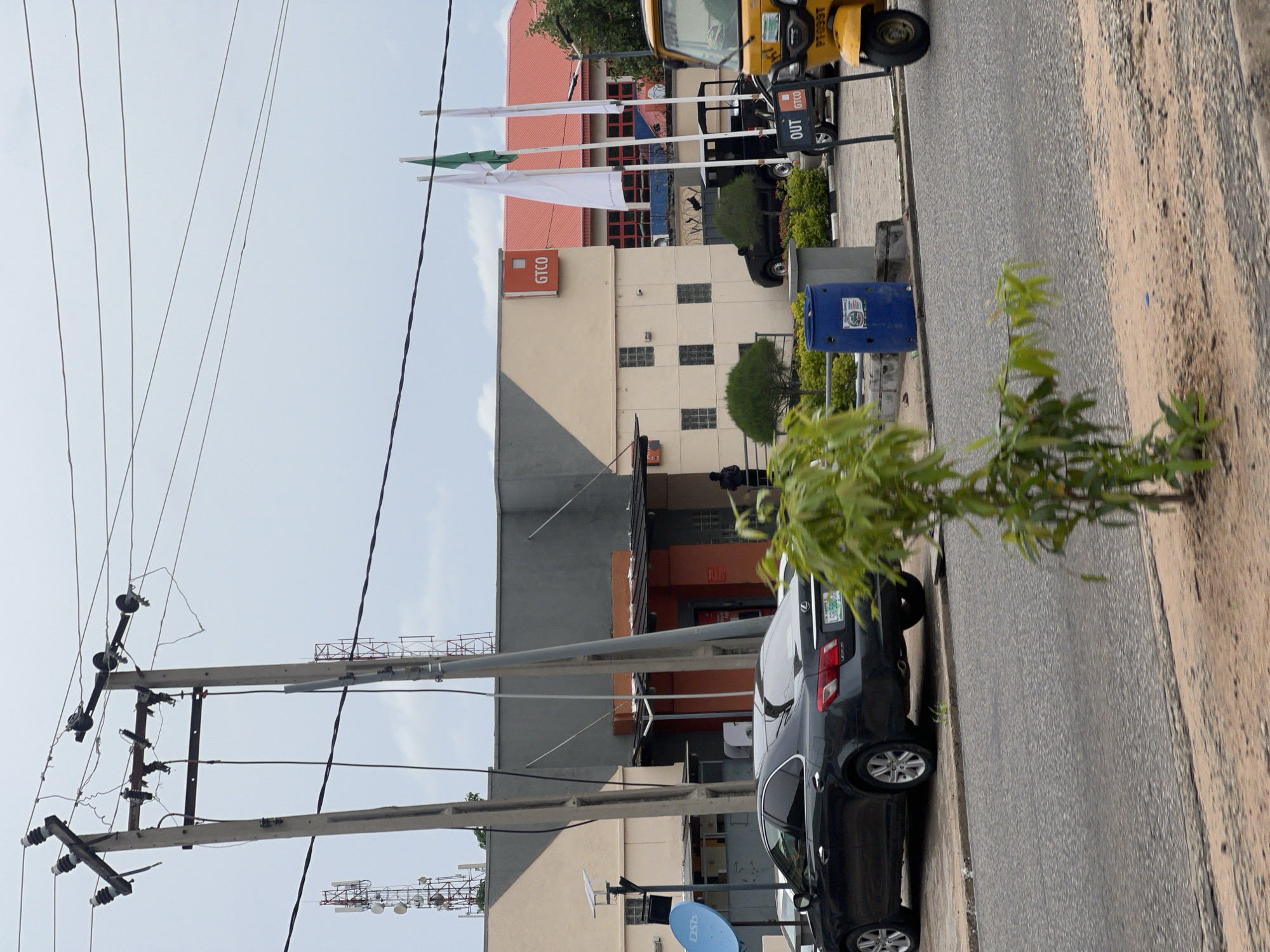 The image depicts a street scene in Jalingo, Nigeria. A dark sedan is parked on the side of the road in front of a building with a GTCO sign. To the right, a yellow three-wheeled vehicle is visible, along with a sign that reads "OUT GTCO". The sky is overcast, and power lines run across the top of the frame, supported by utility poles. A small tree with green leaves is in the foreground, partially obscuring the view. The building appears to be a commercial establishment, possibly a bank, given the signage. The overall environment suggests an urban setting with visible infrastructure and vehicles.
