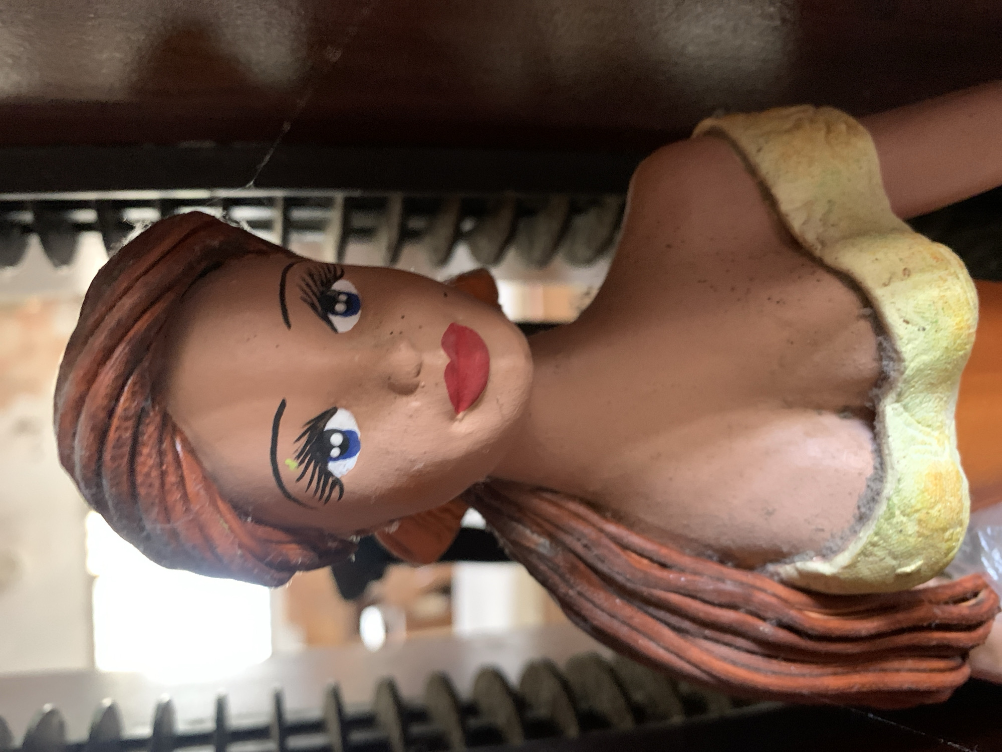This media file captures a close-up of a handcrafted ceramic or terracotta bust of a woman, displayed indoors. The figure is oriented sideways in the frame, depicting her head, shoulders, and upper torso. Her sculpted hair is reddish-brown, styled in flowing waves. The face, with a light complexion, features large, painted blue eyes with long, dark eyelashes and defined eyebrows. Her lips are painted a vibrant red. She is adorned with what appears to be a light yellow, textured garment or top covering her chest and shoulders.
The bust is placed on a dark surface, possibly a wooden shelf or display unit, which is visible in the foreground and background. Behind the figure, a dark, ridged structure, resembling a rack or support, can be seen. In the distant background, a bright, diffuse light source, likely a window, casts a soft glow, suggesting daytime. The image focuses solely on the static object, with no human activity or specific event taking place. The craftsmanship suggests an artisanal piece. There is no visible text in the image.