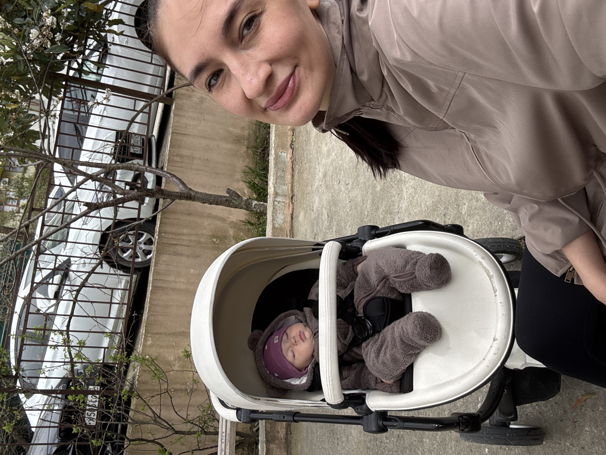 A woman is taking a selfie with a baby in a stroller on a sidewalk in Sochi, Russia. The woman is fair-skinned with brown hair pulled back, wearing a light brown jacket. The baby is asleep in a white stroller, wearing a grey furry outfit and a purple knit cap with ears. Behind them is a white car parked behind a metal fence. A tree with budding leaves is next to the fence. The sidewalk is concrete.
