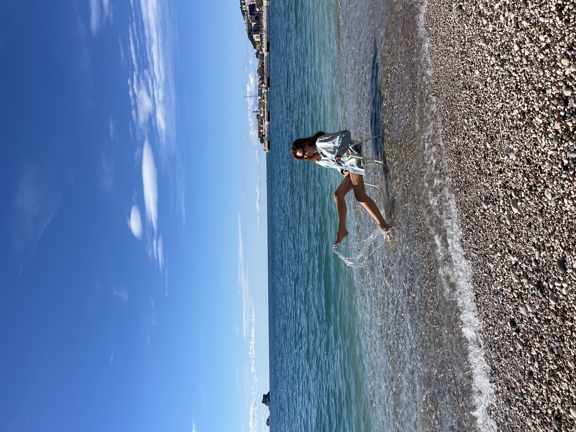 A woman stands on a beach at the waterline in Budva, Montenegro. The sea is calm, and the sky is clear with scattered clouds. The beach is pebbly. The woman is wearing a light-colored denim-like jacket and sunglasses, and is kicking her bare feet in the water. She is next to a small, white, folding chair partially submerged in the water. A building line is visible in the distance across the water.