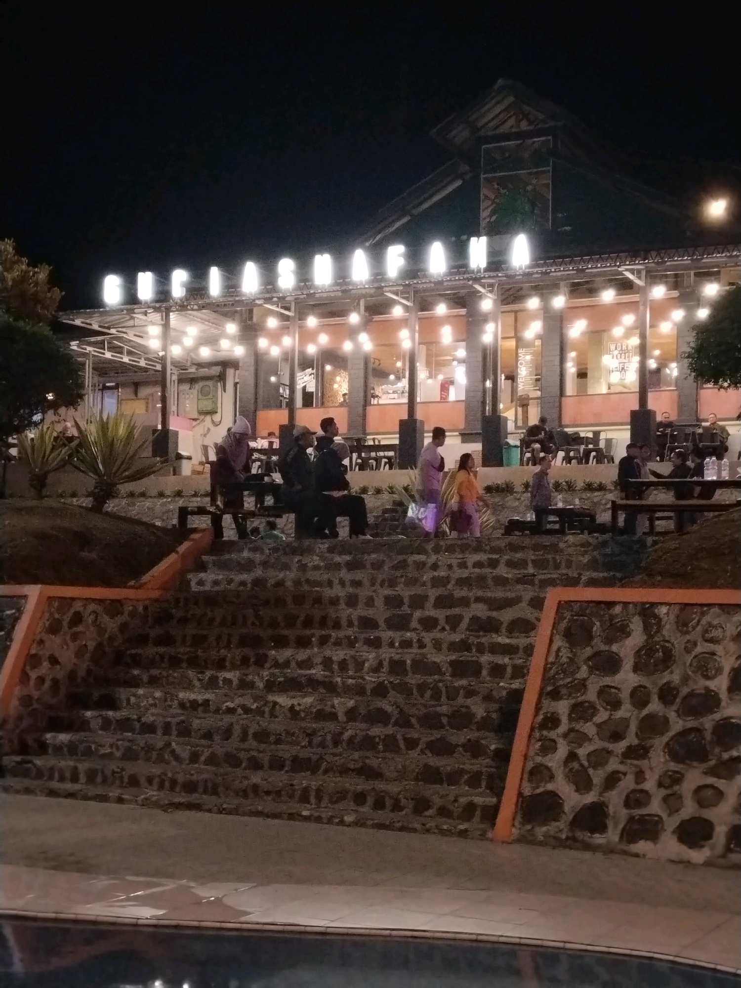 The image captures an outdoor seating area at night, likely a restaurant or cafe, illuminated by numerous string lights and the bright neon sign of "GUCIASAFANA" above the building. The scene is set in Guci, Indonesia, a known tourist destination. In the foreground, a wide stone staircase with orange accents leads up to the main level where people are seated at tables and benches. Several individuals are visible, some standing and others seated, engaged in casual conversation or observing their surroundings. The architecture of the building suggests a modern design with large windows offering a glimpse into the interior. The overall atmosphere appears relaxed and social, typical of a popular evening gathering spot in a tourist area.