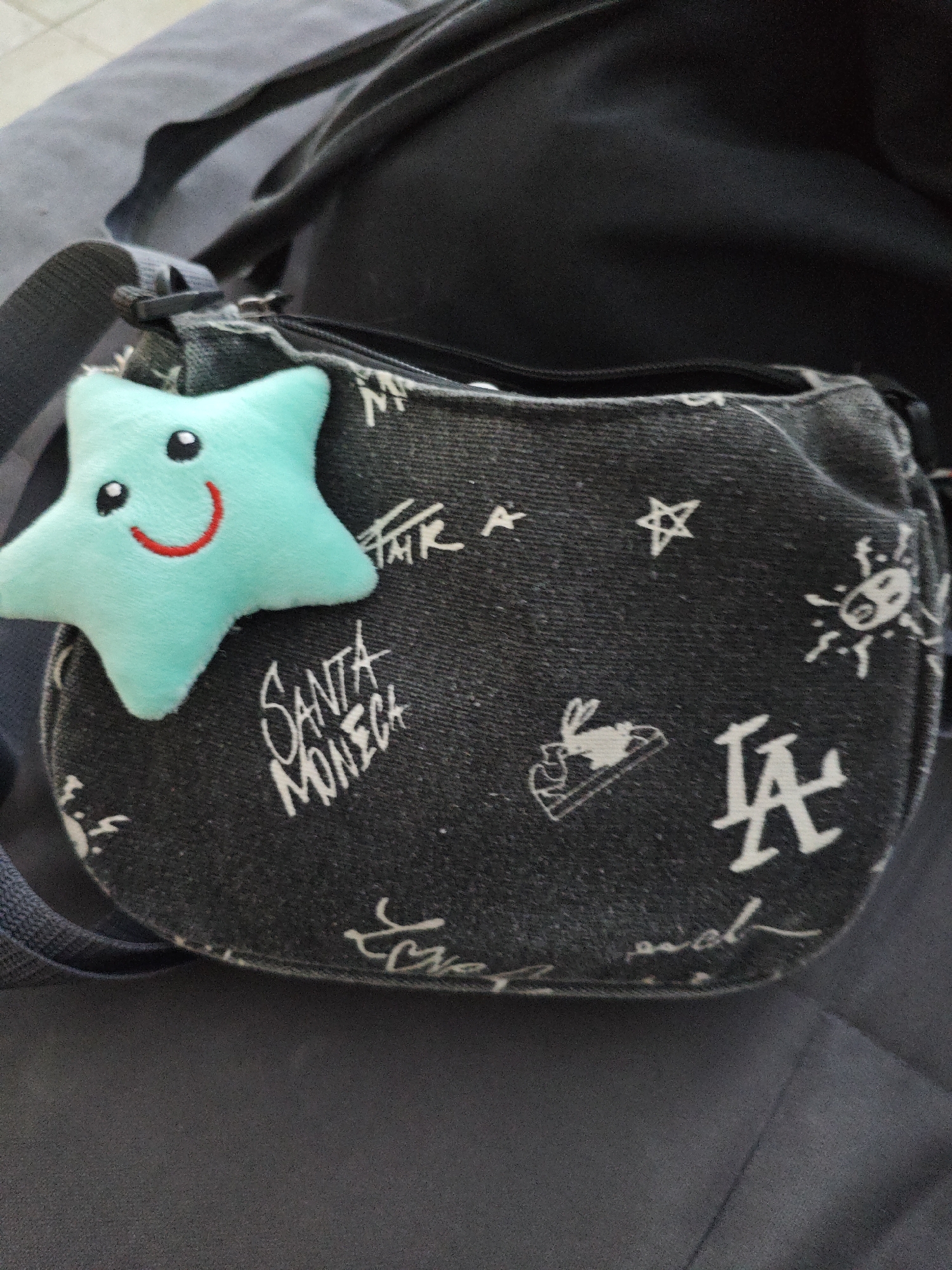 A close-up shot captures a small, teal-colored plush star with black eyes and a red smiling mouth, attached to a dark denim purse. The purse is adorned with various white graffiti-style designs, including the text "SANTA MONICA", a star, a shoe with a rabbit inside, a stylized sun, the letters "LA", and other illegible script. The purse has a zipper closure and a fabric strap. The background consists of a dark gray, textured surface, possibly a piece of furniture. The image is taken from a slightly elevated angle, looking down at the purse and its attached charm. The scene appears to be indoors, with no discernible indication of the specific location within Alushta, Ukraine.