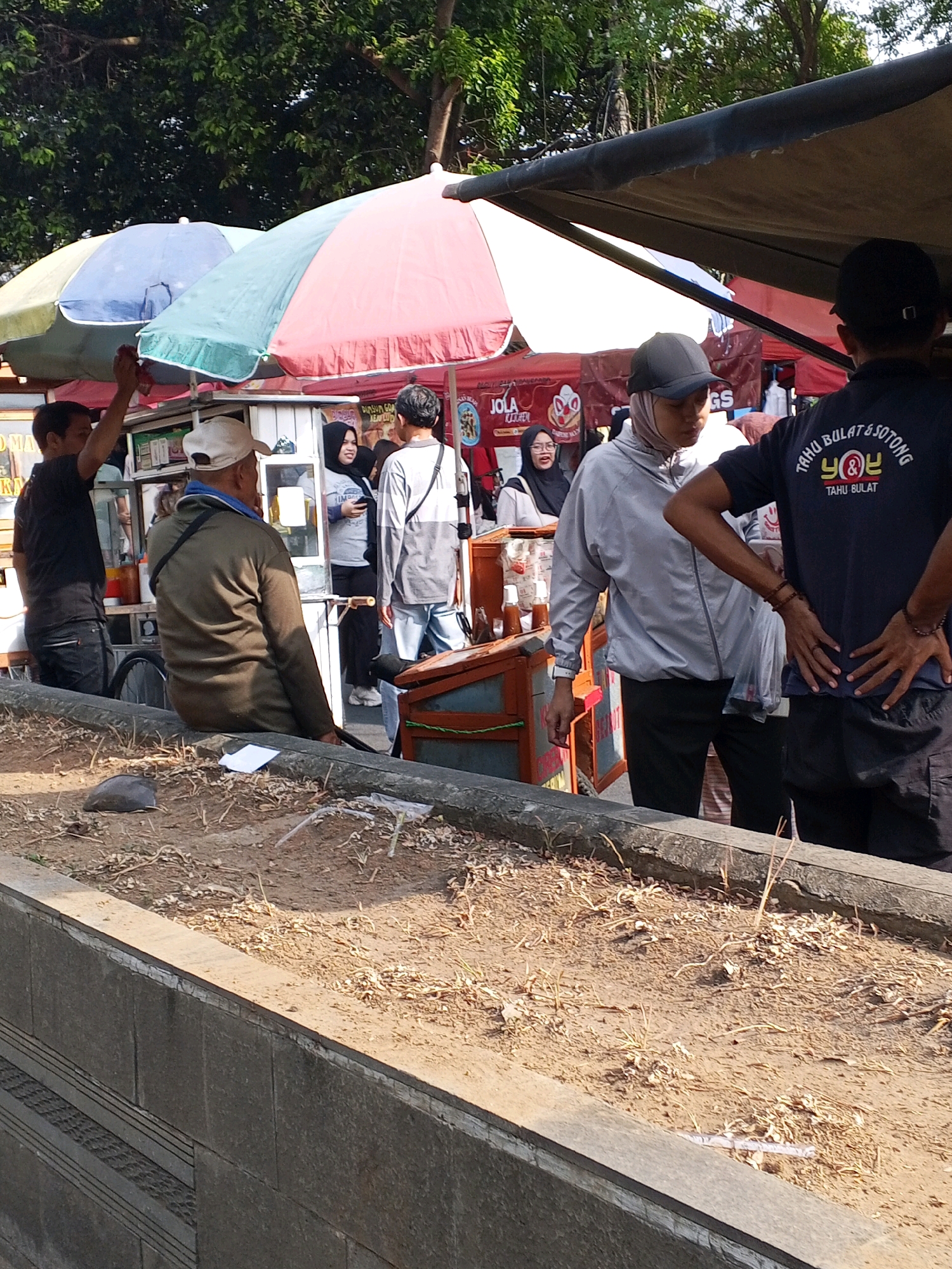 In Serang, Indonesia, a street scene unfolds with several people gathered around food stalls. Underneath colorful umbrellas, two food vendors appear to be serving customers. One vendor, wearing a black t-shirt with Indonesian text and a design, is seen from behind with hands on hips. Another vendor, in a light-colored jacket and headscarf, is facing away from the camera and appears to be interacting with the customer. A third vendor in a brown jacket and cap is seated and looking towards the stalls. The ground in the foreground is dirt with scattered dry leaves and debris, bordered by a low concrete wall. In the background, trees provide a backdrop to the bustling market area.