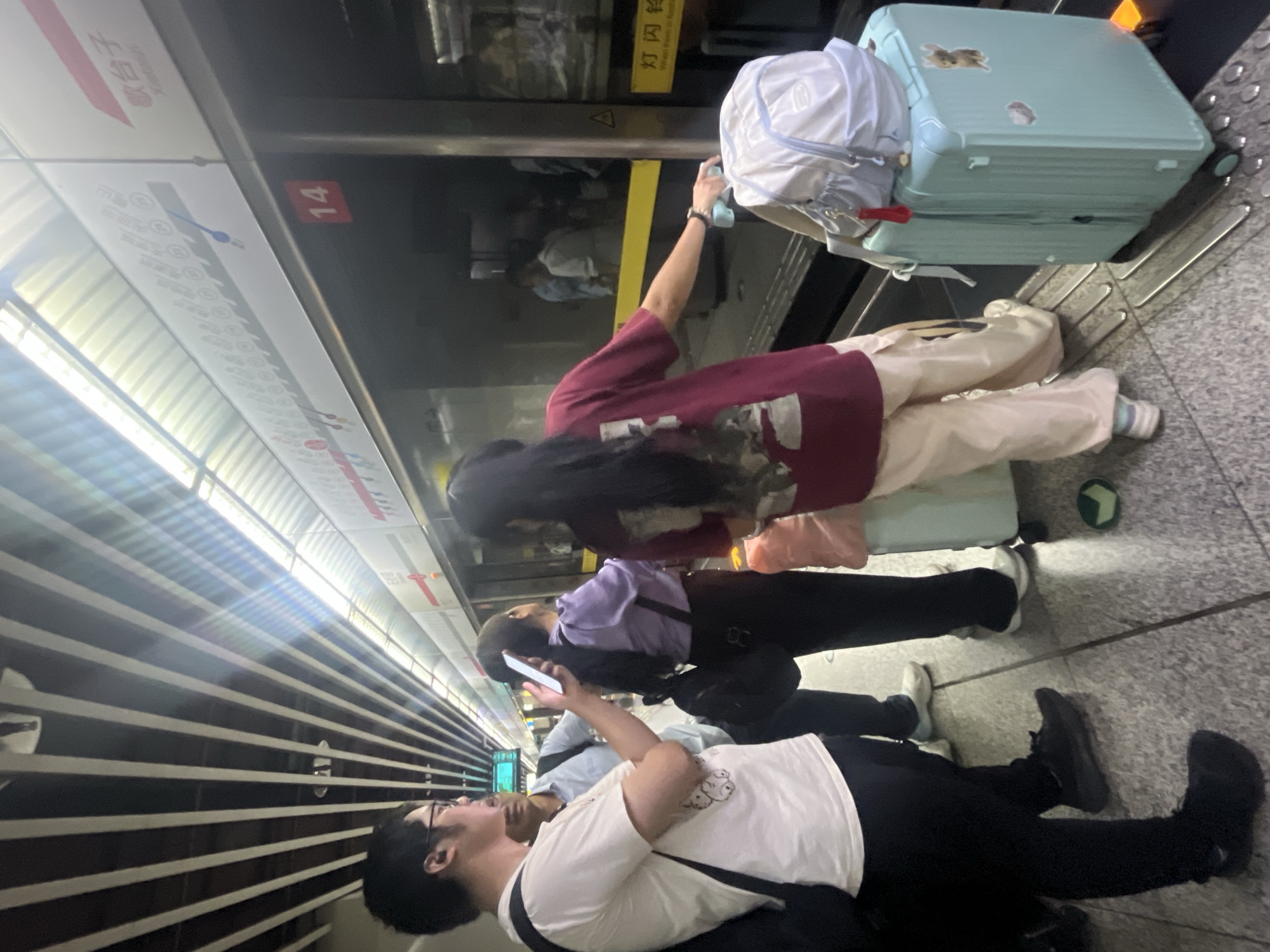 The image depicts a subway platform in Jiulongpo District, China, where several individuals are waiting for a train. The platform features a tiled floor with tactile paving and a yellow safety line along the edge. Overhead, a striped ceiling structure runs the length of the platform, illuminated by artificial lights. A subway train is visible through the glass doors, with its number "14" prominently displayed.
On the platform, a young man wearing glasses and a white t-shirt with a graphic print stands on the left, carrying a black backpack. Beside him, a person in a light blue shirt and dark pants is also waiting. In the center, a young woman in a purple top and dark pants holds a smartphone. To her right, another woman in a maroon t-shirt and light-colored pants is holding onto the handle of a light blue suitcase, which is positioned next to a matching backpack. The woman's hand is near a yellow safety strip on the train door. Further in the background, other passengers are visible inside the train and on the platform. Signs with Chinese characters are present on the wall above the train doors. The overall atmosphere suggests a typical scene of urban public transportation.