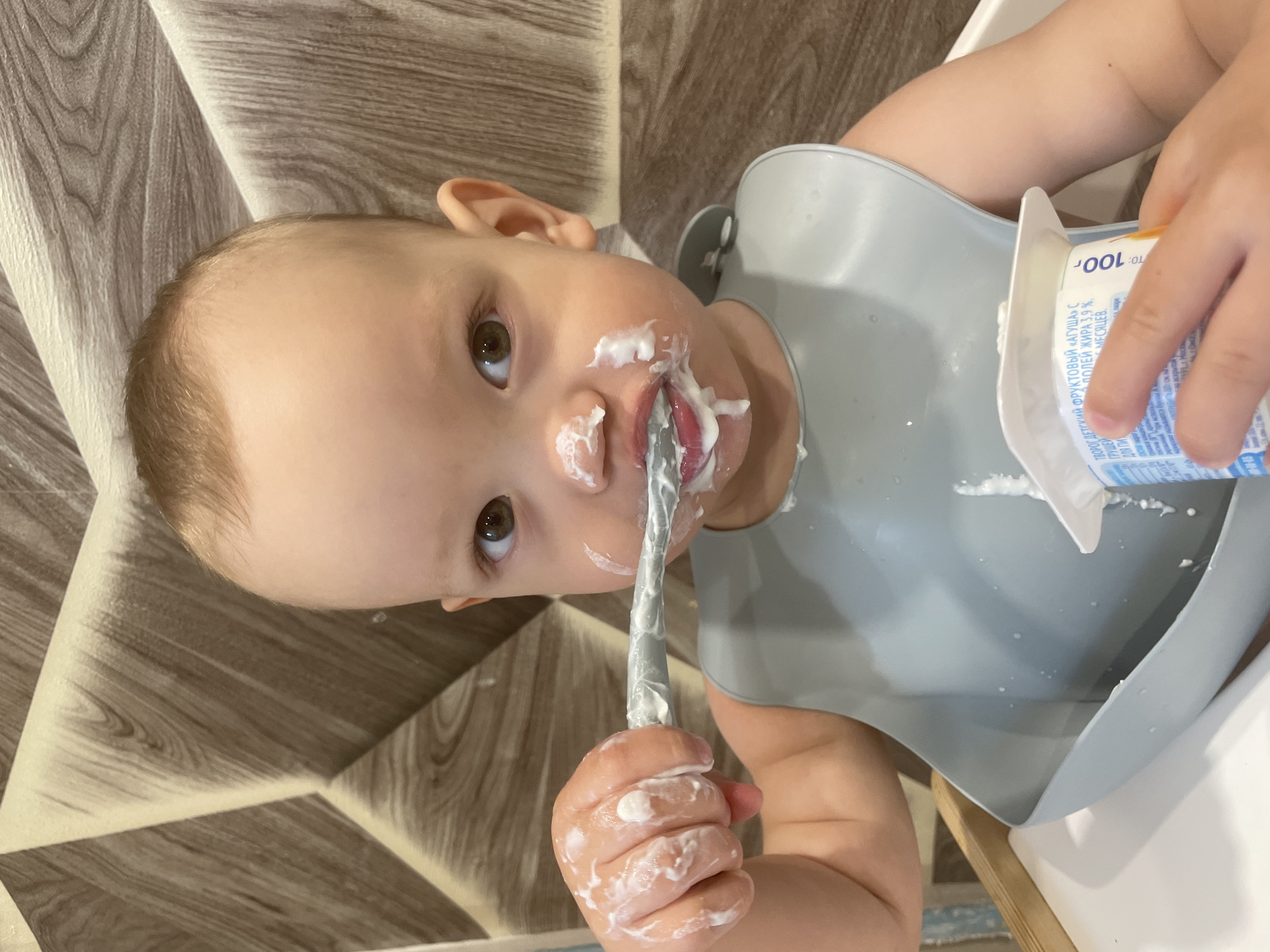 A young child is eating yogurt. The child, wearing a gray bib, holds a spoon to their mouth. Yogurt is smeared around the child's mouth and on their hand. A container of "Agusha" fruit yogurt is visible in the upper right, printed in Russian. The container indicates that the yogurt is pear-flavored, with 3.9% fat, and is intended for children of a certain age. The background shows a wood-patterned surface. The picture does not contain enough information to identify the exact location in Perm, Russia.