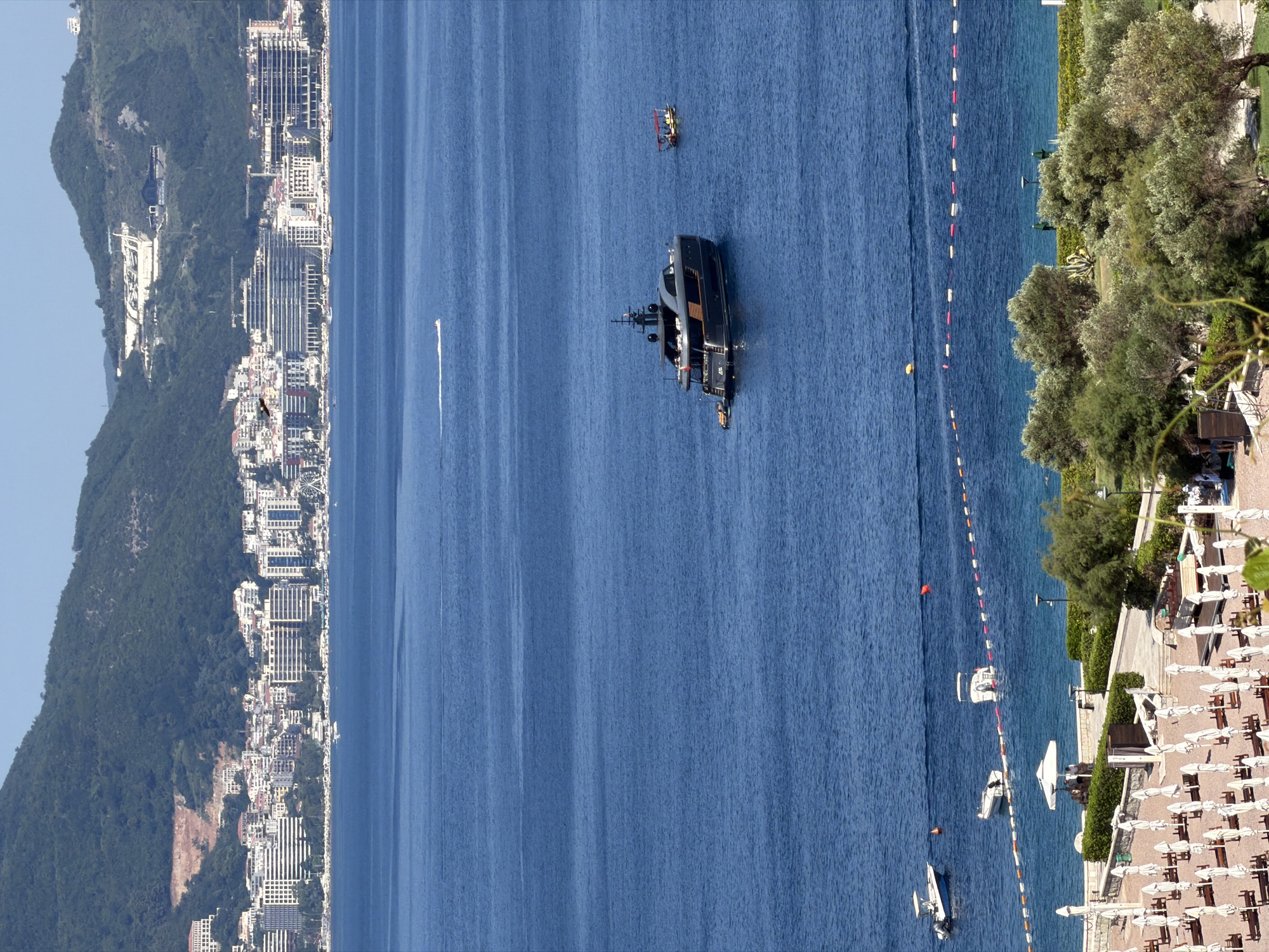 This is an aerial, wide shot of the coastline in Sveti Stefan, Montenegro, on a clear, sunny day. The foreground features a resort area with numerous white beach umbrellas and lounge chairs neatly arranged on a sandy beach. Beyond the beach is the blue Adriatic Sea, marked by a floating barrier of red and white buoys. Several small white boats are visible in the water near the shore, with one further out and partially submerged. In the middle ground, a large, dark-colored yacht is anchored. To the right of the yacht, a small, red-roofed boat is also present. In the background, a densely populated city with numerous white buildings stretches along the coast at the base of a steep, green, tree-covered mountain. The mountain summit is capped by a light-colored, multi-level structure. The sky is a clear, bright blue.