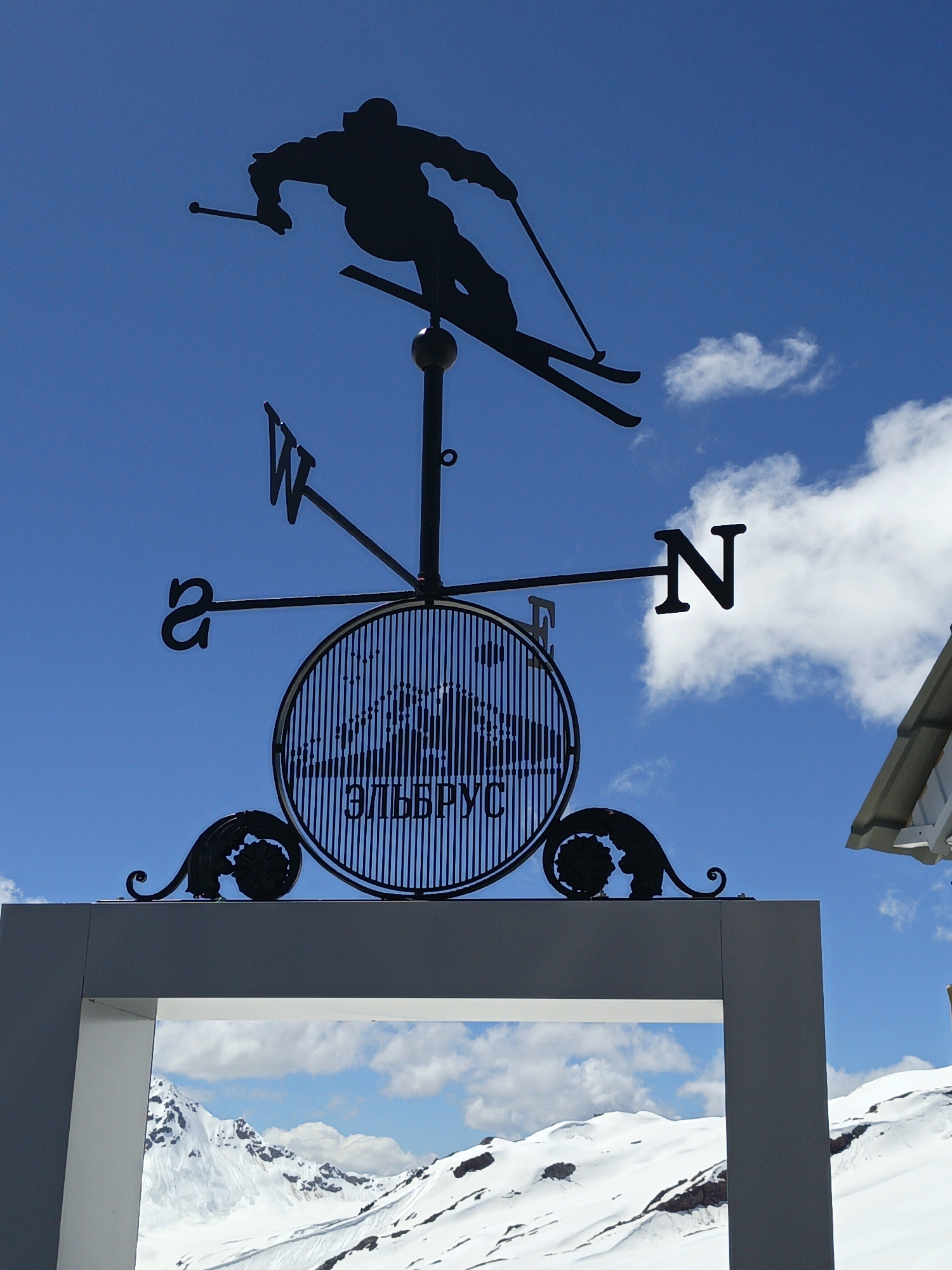 A black silhouette of a skier mid-jump is mounted on a weathervane against a bright blue sky with scattered white clouds. The skier's skis are angled downwards and to the right, and they hold ski poles. The weathervane also features directional indicators: "W" to the left, "N" to the right, and partially visible "S" to the bottom left. A circular ornament with vertical lines and the Cyrillic text "ЭЛЬБРУС" (Elbrus) is centrally located below the skier. Decorative black metal scrollwork is visible on either side of the circular ornament. The weathervane is mounted on a black pole. Below the weathervane, a portion of a modern, grey-framed structure is visible, framing a snow-covered mountain landscape. The mountains are white and rugged, with some exposed rock and patches of darker snow or ice. This scene is located in Terskol, Russia, likely at a viewpoint or resort area, given the prominent weathervane and the view of the snow-capped mountains.