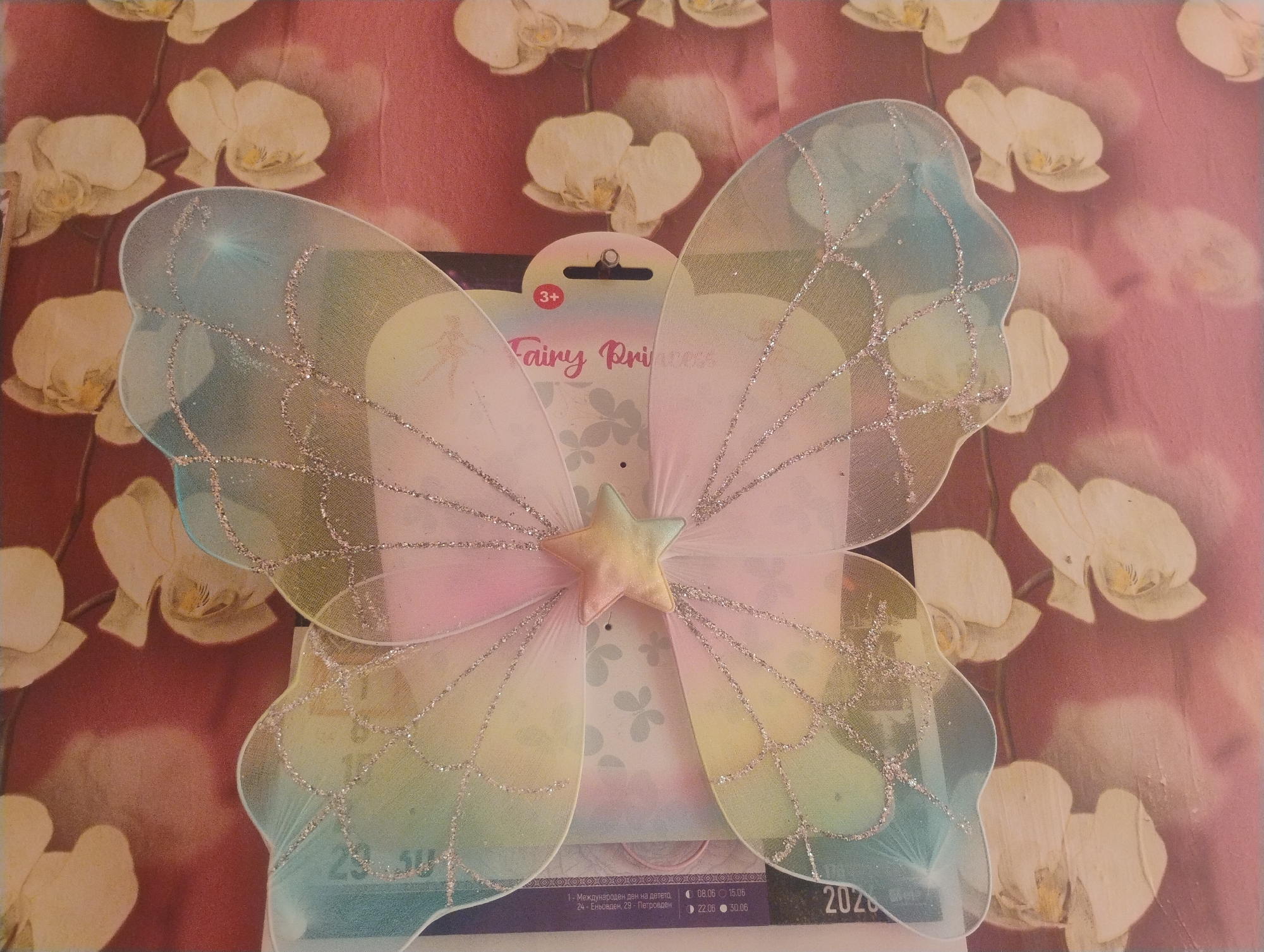 The image shows a pair of iridescent fairy wings with glitter accents, attached to a cardboard backing that reads "Fairy Princess" and has a "3+" age recommendation. The wings are translucent and feature a gradient of pastel colors including pink, yellow, and blue, with a star embellishment at the center. The background is a fabric with a floral pattern of white orchids against a pink and burgundy gradient. The image is a top-down view of the product. There are no people or actions depicted. The location is not specified in the image itself.