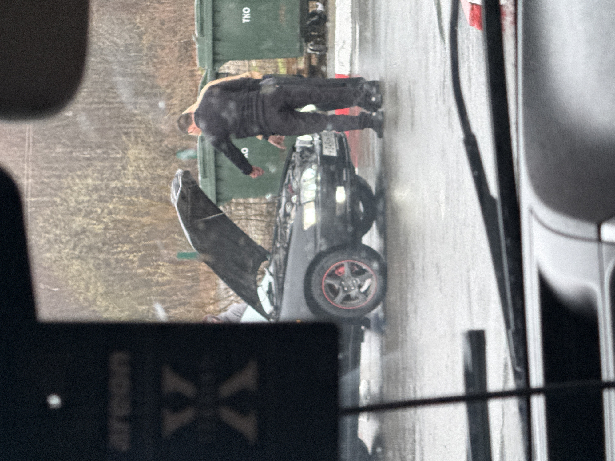 The image captures an outdoor scene, likely a street or parking area, under overcast daylight conditions, indicated by the wet ground. The primary subject is a man, dressed in dark clothing (black long-sleeved shirt and pants), standing beside a black car with its hood wide open. He appears to be leaning over, either inspecting or working on the car's engine.
In the background, two dark green dumpsters or waste bins are visible. One dumpster clearly displays the text "TKO" on its side. Beyond the bins, out-of-focus trees or brush create a natural backdrop.
The car itself is black, and one of its wheels, visible in the foreground, features a distinctive red accent on its rim. The overall setting suggests a car breakdown or maintenance in progress.
In the immediate foreground, a blurred object obstructs part of the view. Upon closer inspection, the text "Areeon XX" is visible on this object, which appears to be an air freshener, implying the photo was taken from inside another vehicle. The ground is consistently wet, suggesting recent rainfall. No strong emotional cues are evident from the man's posture, which indicates focus on the task.