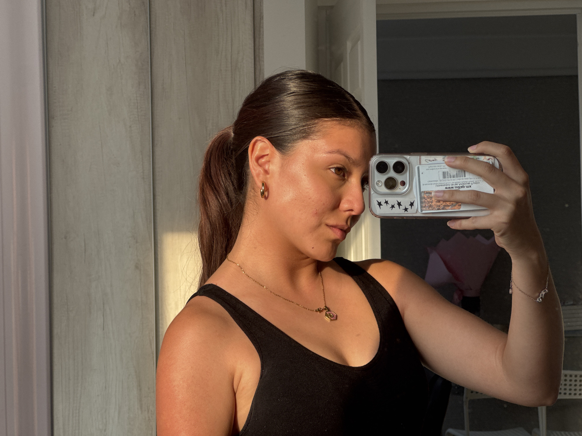 A young woman is taking a selfie indoors. She has dark hair pulled back in a ponytail, gold hoop earrings, and a gold necklace with a pendant. She is wearing a black tank top. Her phone has a clear case with star stickers and other items tucked inside. The background includes a textured wall and a doorway leading to a brighter area. Reflections in the glass door suggest furniture and potentially a bouquet of flowers. It is difficult to confirm the exact location but there is no reason to believe it is in San Francisco Ocotlán (Ocotlán), Mexico.