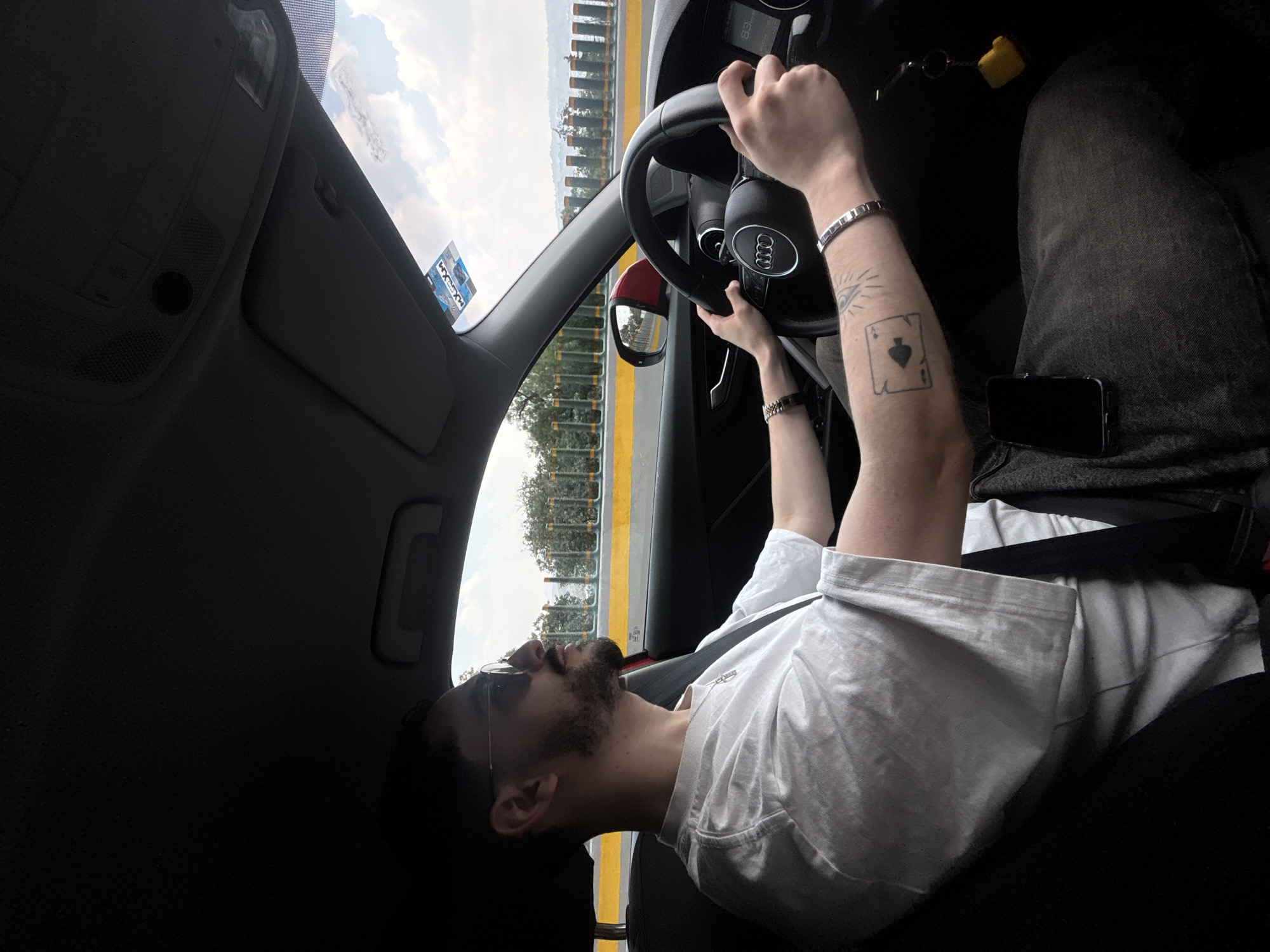 A person is seated in the driver's seat of an Audi automobile. The person is wearing a white t-shirt, sunglasses, and has a beard. The person has an Ace of Spades tattoo on their left forearm, a silver bracelet on their left wrist, and a watch on their right wrist. The car is on a road with a yellow and green barrier visible through the window. A mobile phone is on the person's right leg. The location of the scene is Unknown, Unknown.