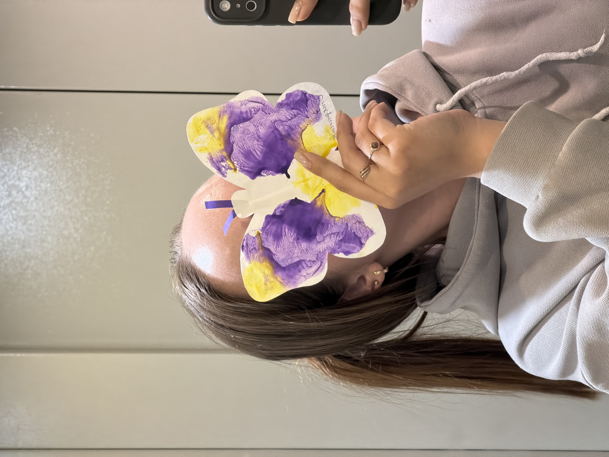 A person is holding a paper butterfly with purple and yellow paint in front of their face, presumably to conceal it. The person has long brown hair and is wearing a light gray hooded sweatshirt. They are also wearing rings on their fingers. The background appears to be a plain white or light-colored wall. The photo is taken with a mobile phone. There is no indication of the location as Chaikovsky, Russia.