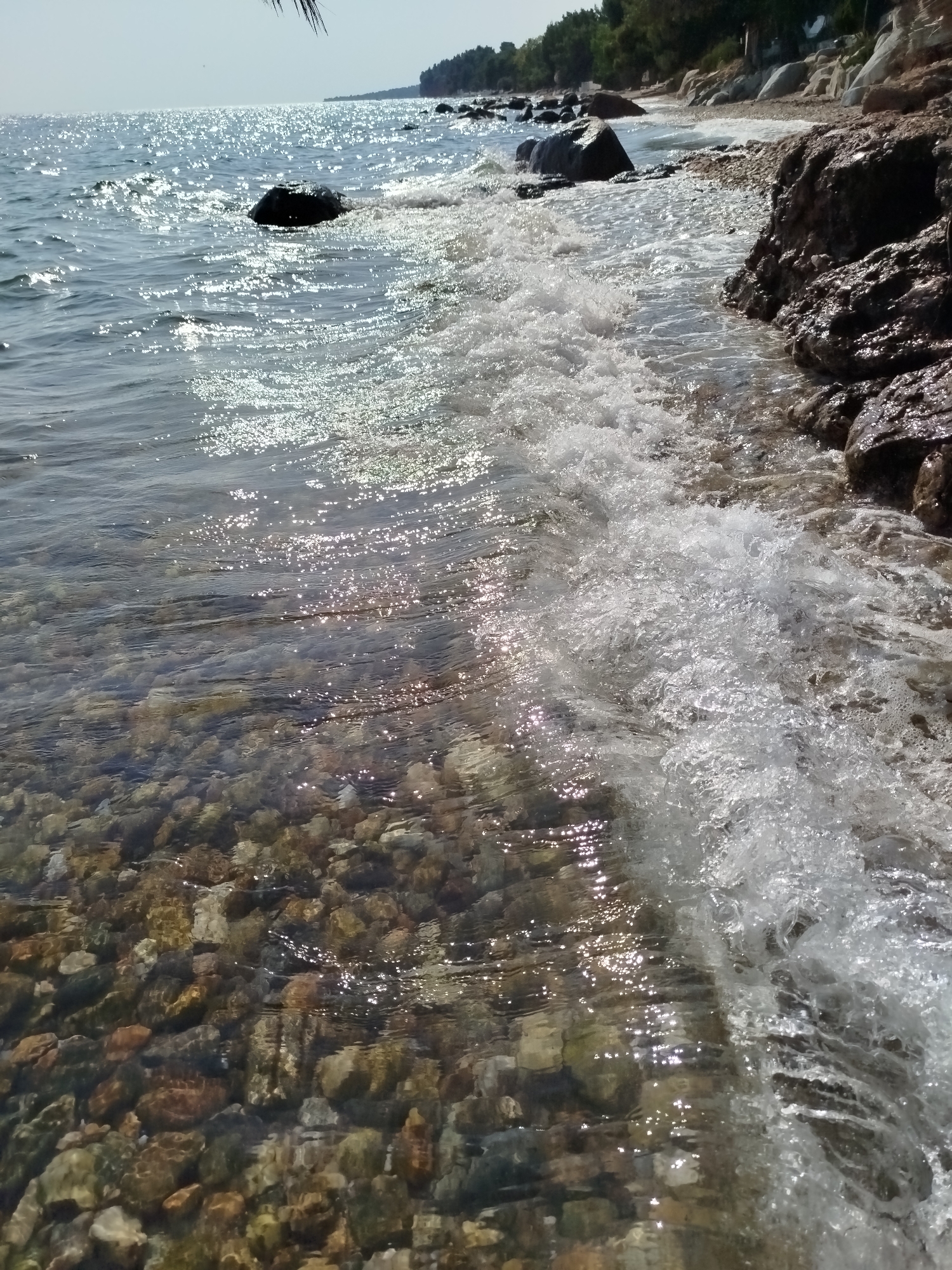 This image captures a sunlit beach scene in Sithonia Municipal Unit, Greece. The foreground is dominated by clear, shallow water revealing a bed of colorful pebbles. Small waves gently lap the shore, creating foamy white water and shimmering light reflections on the surface. The shoreline is rocky, with larger, dark stones visible on the right side of the frame. In the middle ground, larger dark rocks are partially submerged in the water. The background shows a more distant shoreline with trees and a hint of a clear sky. No people are visible in the image.