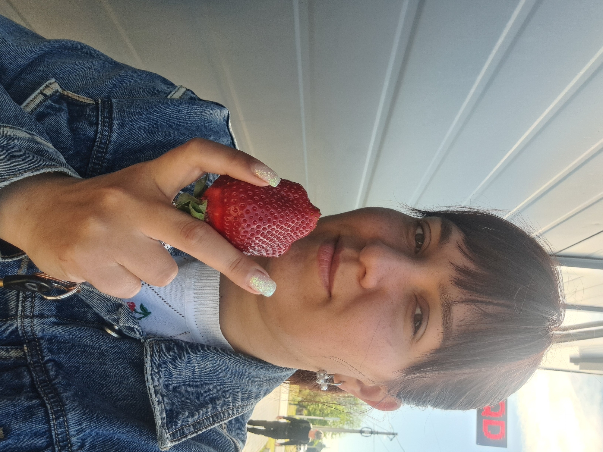 A woman is holding a strawberry up to her face. She is wearing a blue denim jacket over a white shirt with an embroidered flower on the collar. She has short dark hair and a small silver earring. Her nails are painted with glitter polish. She is holding keys in her left hand. The background includes a white paneled wall and a street scene. A traffic light with a red light is visible, as well as two figures walking in the distance. The scene appears to be Leningradskaya, Russia.