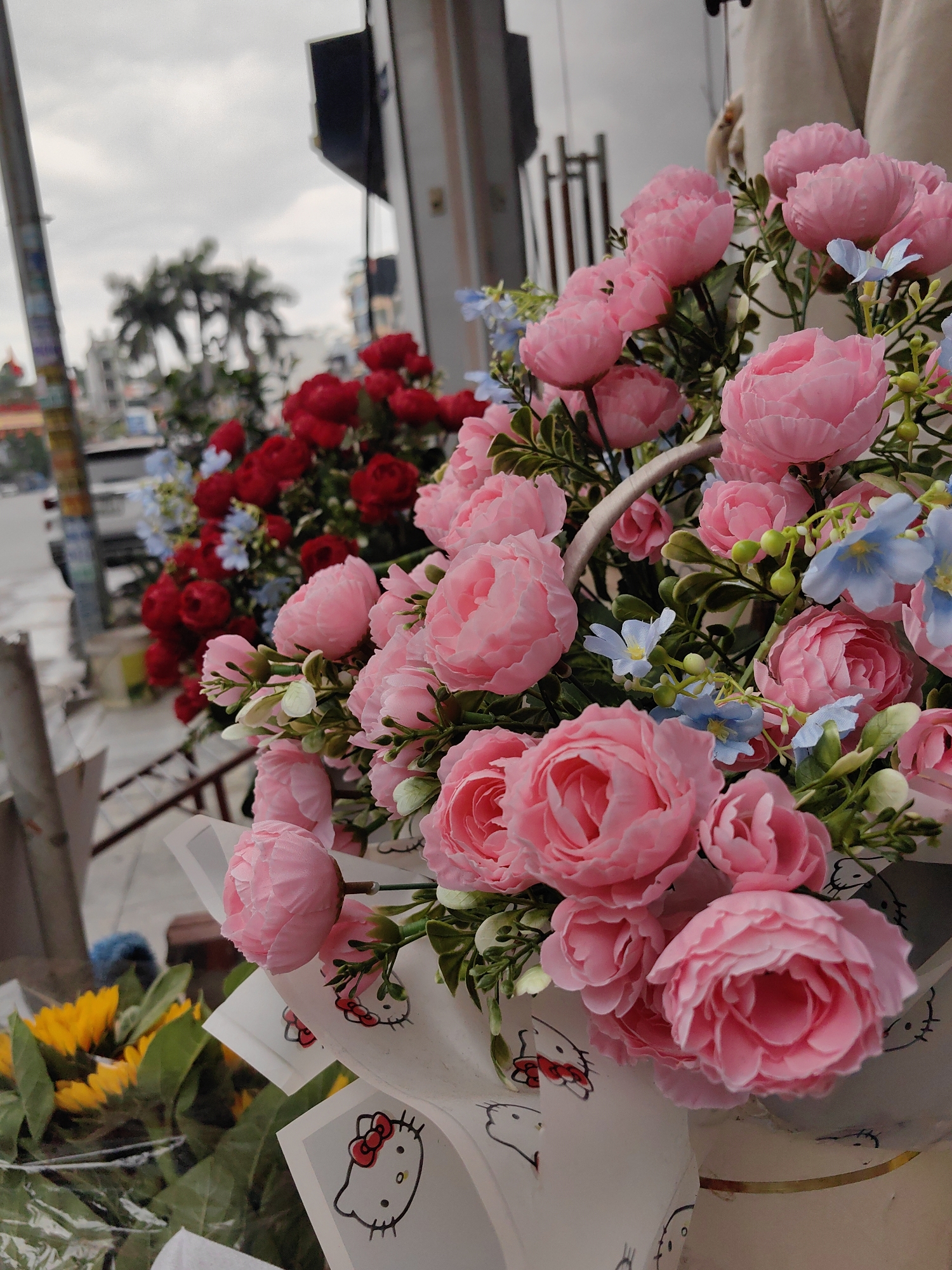 The image shows a display of artificial flower bouquets for sale, likely on a street in Bai Chay Ward, Vietnam. The focus is on a bouquet of pink peony-like flowers interspersed with smaller blue flowers, wrapped in paper featuring a Hello Kitty design. To the left are bouquets of red flowers and a hint of sunflowers. In the blurred background, there is a street with buildings, utility poles, palm trees, and vehicles, suggesting an urban setting on an overcast day. The light is diffused, creating a soft, muted color palette. The bouquets appear to be positioned on a ledge or shelf, possibly outside a shop.