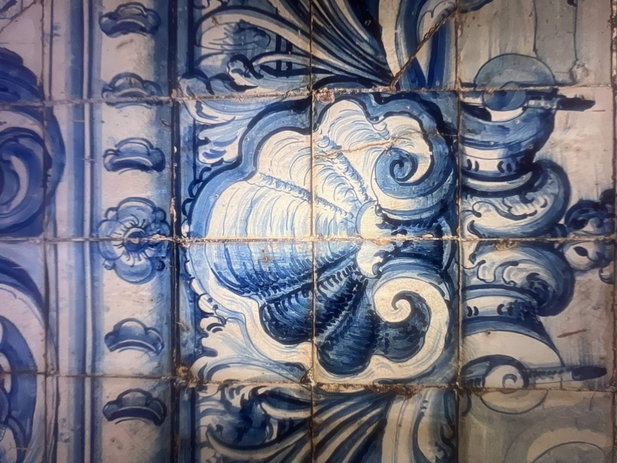 The media file presents a close-up view of a wall adorned with traditional blue and white ceramic tiles, known as azulejos, originating from Mollet Del Vallès, Spain. The subjects are the individual tiles forming a larger decorative pattern.
The tiles feature intricate, hand-painted designs in various shades of blue, from deep indigo to lighter tones, set against a creamy white background. The central pattern is characterized by ornate, curvilinear motifs, including large, stylized scrolls, elaborate foliage, and what appear to be shell-like forms or architectural elements resembling parts of a column or capital with volutes. These designs create a dynamic and flowing composition across the tiled surface. On the vertical sections, smaller, repeating cloud-like or stylized floral elements form a border.
No activity or event is taking place, as the image focuses solely on the static architectural decoration. Notable details include the clear visibility of the grout lines between the tiles, indicating their individual placement and construction. The tiles exhibit signs of age and authenticity, such as minor imperfections, slight wear, and subtle variations in color and texture, consistent with traditional craftsmanship. The lighting in the image highlights the glazed surface and the tactile quality of the painted details. There is no visible text on the tiles. The overall scene conveys a sense of historical artistry and decorative tradition prevalent in Spanish architecture.