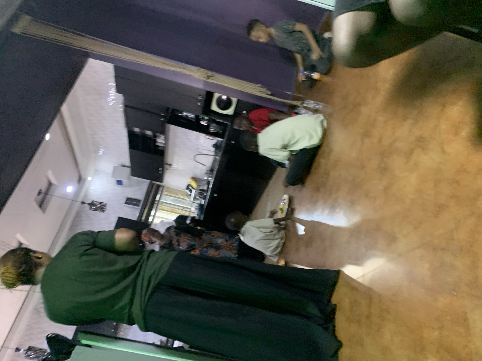 The image depicts an indoor residential scene in Jos, Nigeria, featuring multiple individuals within a living area integrated with a kitchen. In the foreground, a person with blonde hair styled in an updo wears a dark green short-sleeved top and dark, wide-leg trousers, standing with their back to the camera. Behind this individual, a woman in a multi-colored patterned dress stands near a modern kitchen equipped with dark-colored upper and lower cabinets, a stainless steel sink with a faucet, and a white tiled backsplash.
On the light brown, glossy floor, four children are seated. To the left, a child in a light-colored shirt and dark trousers is leaning forward, seemingly interacting with food on a white oval plate. Further back, another child in a red shirt and a third child in a light-colored shirt are seated on the floor. To the right, a fourth child, wearing a dark patterned shirt, sits with legs crossed, with plastic water and orange beverage bottles placed beside them. The lower legs of an additional person in dark shorts are partially visible in the bottom right corner of the frame. The room features a white ceiling with recessed lighting and two dark pendant light fixtures. A dark purple wall is visible on the right, contrasted with a white textured wall above the kitchen area.