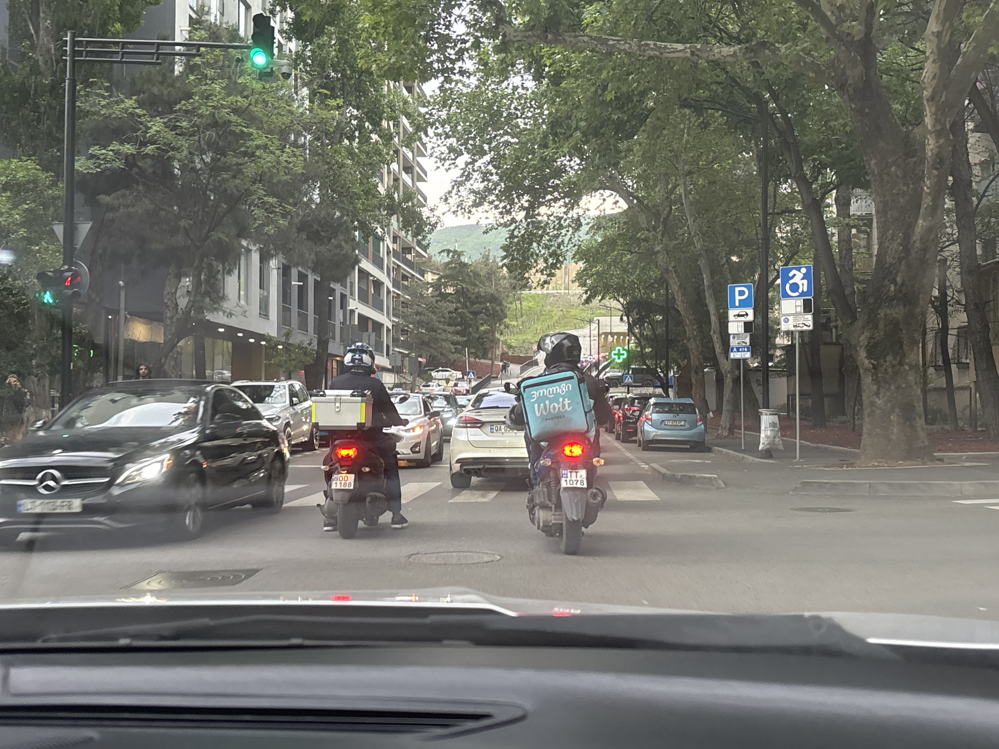 The image shows a street scene in Tbilisi, Georgia, viewed from inside a car. The camera is positioned behind the windshield. Several cars and two motorcycles are in the traffic lane. The motorcycle rider in the foreground has a light-blue "Wolt" delivery backpack. The license plate on the motorcycle is "TT 1078." To the left, a black Mercedes sedan is also visible. A green traffic light signal can be seen in the upper-left. "Parking" and "Handicapped" road signs are on the right. Trees line the street and buildings are on both sides. A mountain range is visible in the far distance down the street.