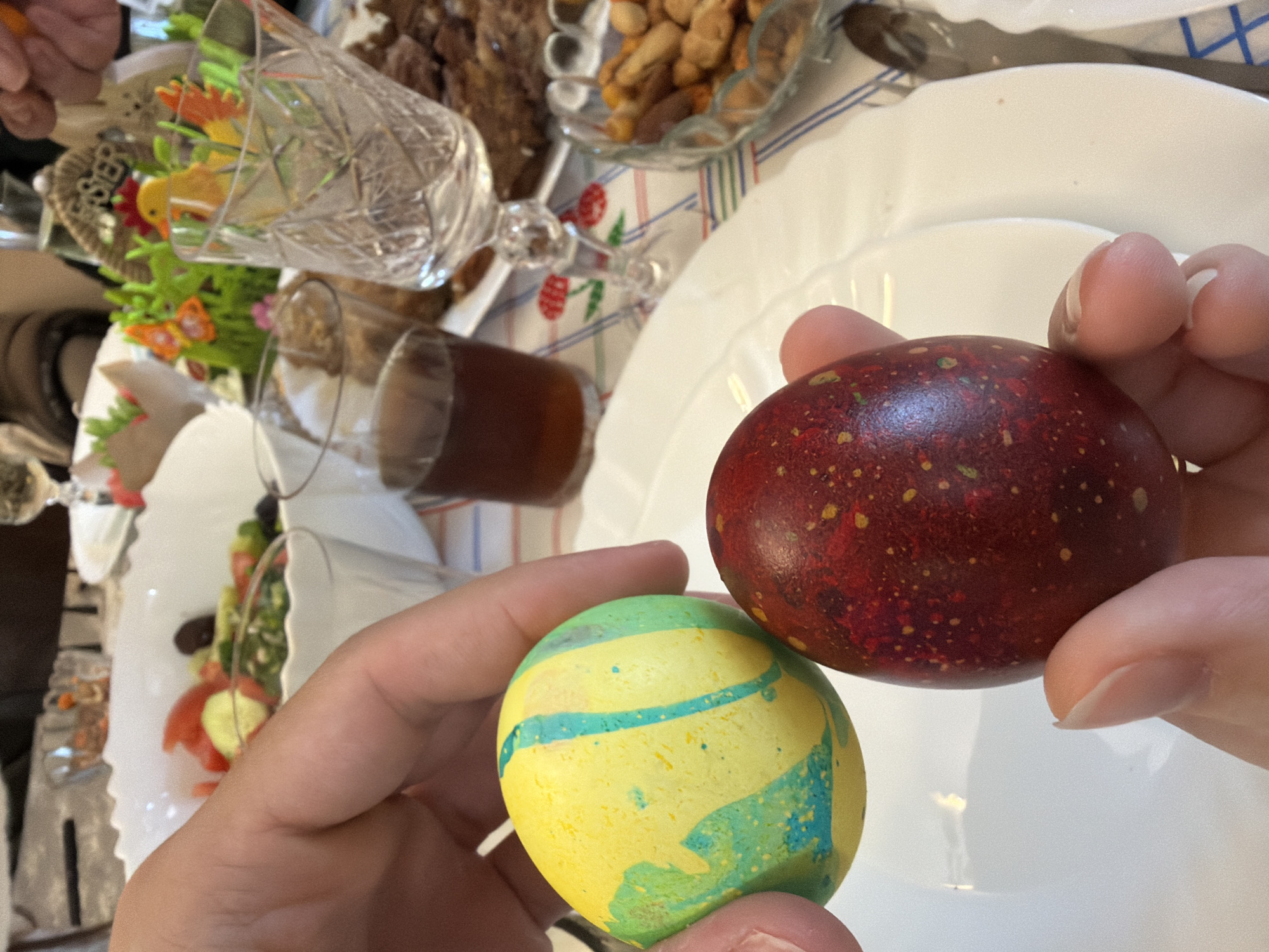The image shows a close-up of two hands holding two dyed eggs, one red with yellow speckles and the other yellow with blue and green swirls. These are positioned over a white plate. The surrounding table setting is visible, featuring a glass, food in bowls, and a salad in a white serving dish. While the location is stated to be Plovdiv, Bulgaria, there are no discernible elements within the image to confirm this.