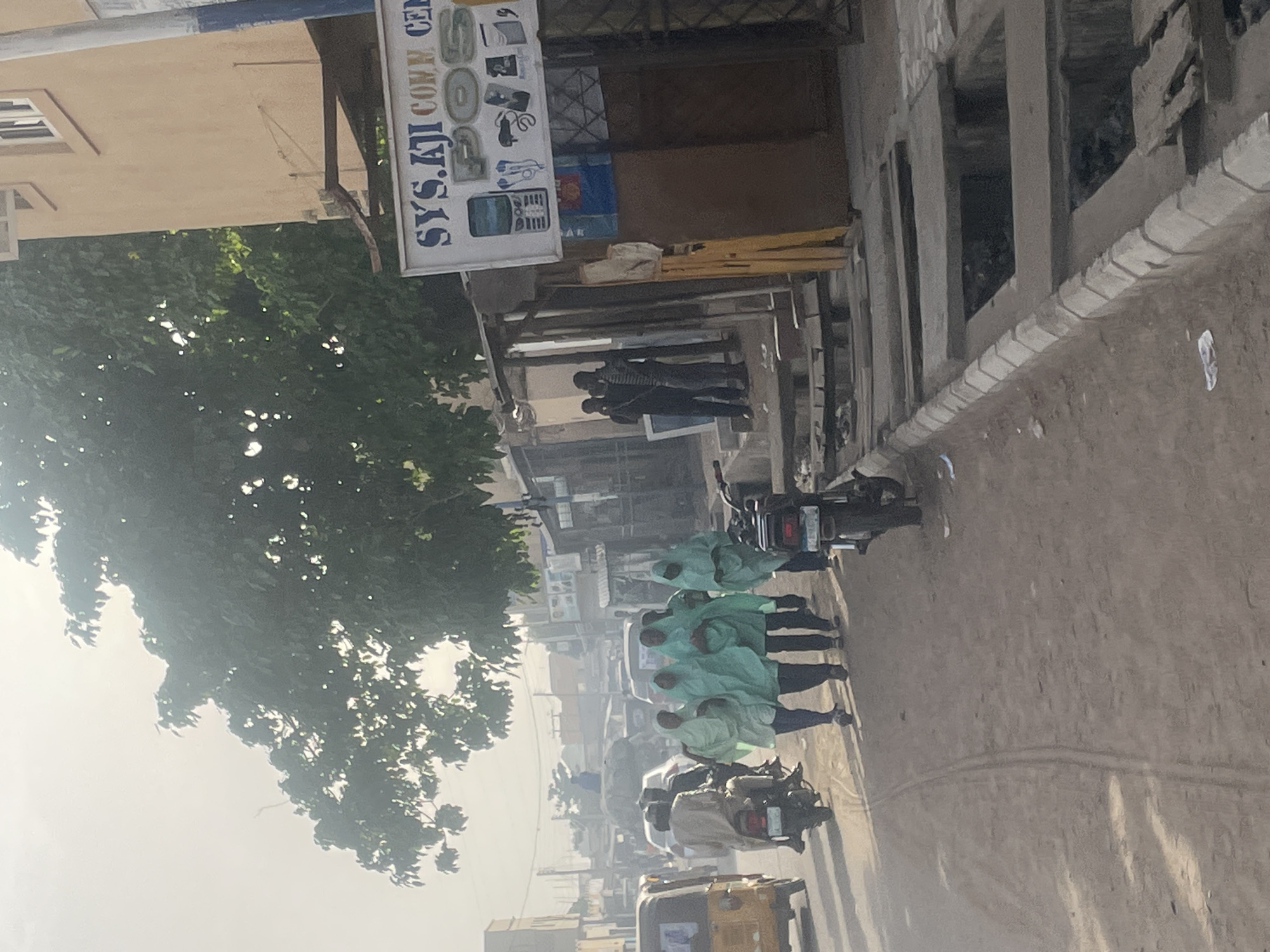 The image depicts a hazy daytime street scene in an urban or semi-urban environment. The perspective is from slightly above, looking down a dusty or unpaved road.
In the mid-ground on the right side of the street, a group of at least four individuals is walking. They are prominently dressed in identical light green or teal hooded garments that cover most of their bodies. A motorcycle is parked close to them. Further down the road, another motorcycle is visible, and in the bottom-left corner, part of a yellow auto-rickshaw can be seen.
On the left side of the frame, a large tree with dense green foliage fills a significant portion of the view. Behind the tree, a building features a visible sign that reads "SYS.AJI COMM. CEN," likely standing for "SYSTEM AJI COMMUNICATION CENTER." The sign also displays images of mobile phones, chargers, and other electronic devices. Two individuals are standing near the entrance of this establishment.
Opposite, on the right side of the street, another building with an unfinished or derelict concrete structure and multiple openings lines the thoroughfare. The overall atmosphere is marked by a strong atmospheric haze or dust, which diffuses the sunlight and blurs details in the distance, suggesting a dry and possibly warm climate. The ground appears uneven and unpaved in sections, indicating regular foot and vehicle traffic.