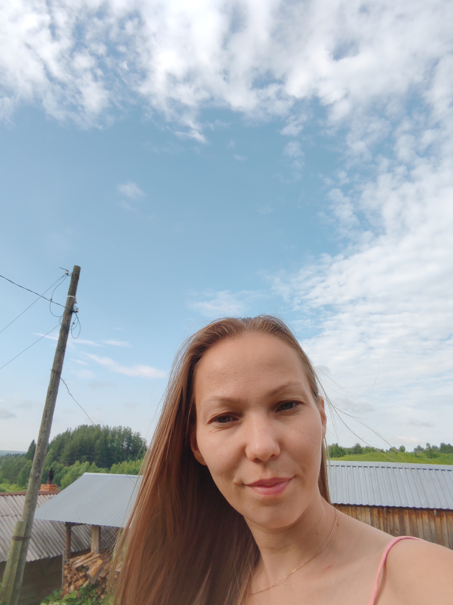 A blonde woman takes a selfie outdoors with a blue sky and white clouds above. She is wearing a pink top and a thin gold necklace. To her left is a wooden utility pole with wires. In the background, there are several wooden structures with corrugated metal roofs, a chimney, piles of firewood, and a densely wooded area. The scene appears to be in a rural or village setting, potentially in Чухлэм, Russia, given the architectural style and vegetation. The lighting suggests it is daytime, likely late morning or early afternoon.