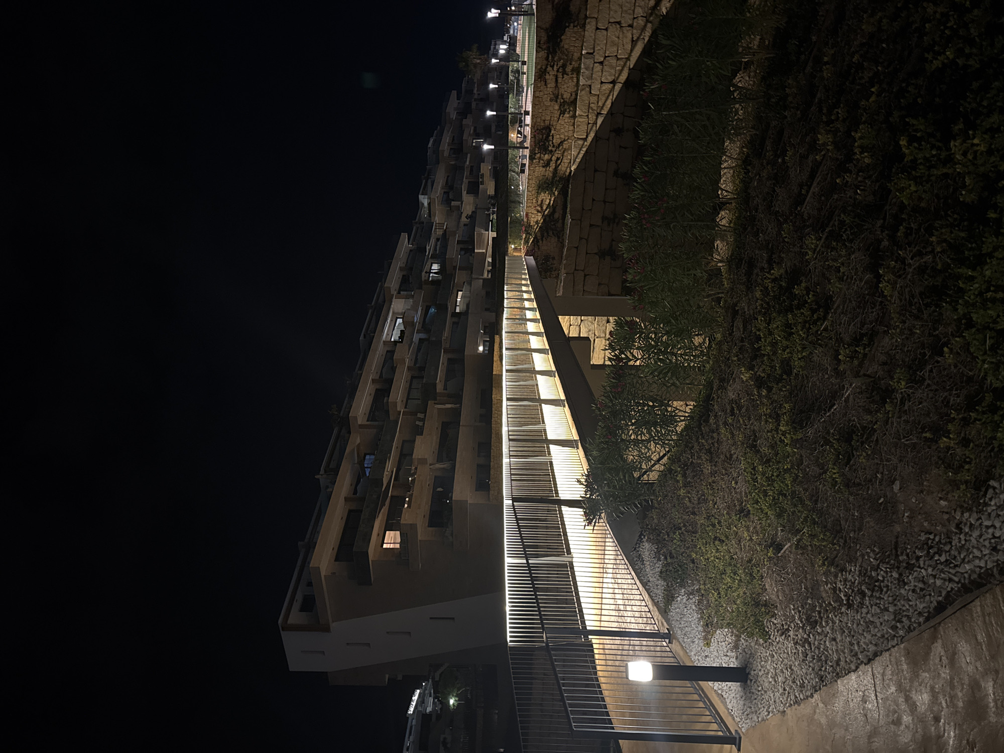 The image depicts a modern, multi-level building with numerous balconies, illuminated at night. In the foreground, a pedestrian bridge with integrated lighting runs diagonally across the scene, leading towards the building. The bridge is bordered by a metal railing and low-lying plants and gravel. A solitary modern lamppost stands to the left of the bridge. The architecture suggests a residential complex, possibly part of a resort or development, situated in a coastal area given the hints of greenery and the style of construction. The overall impression is one of contemporary design and nighttime ambiance. This scene is located in the city of Finestrat, Spain.