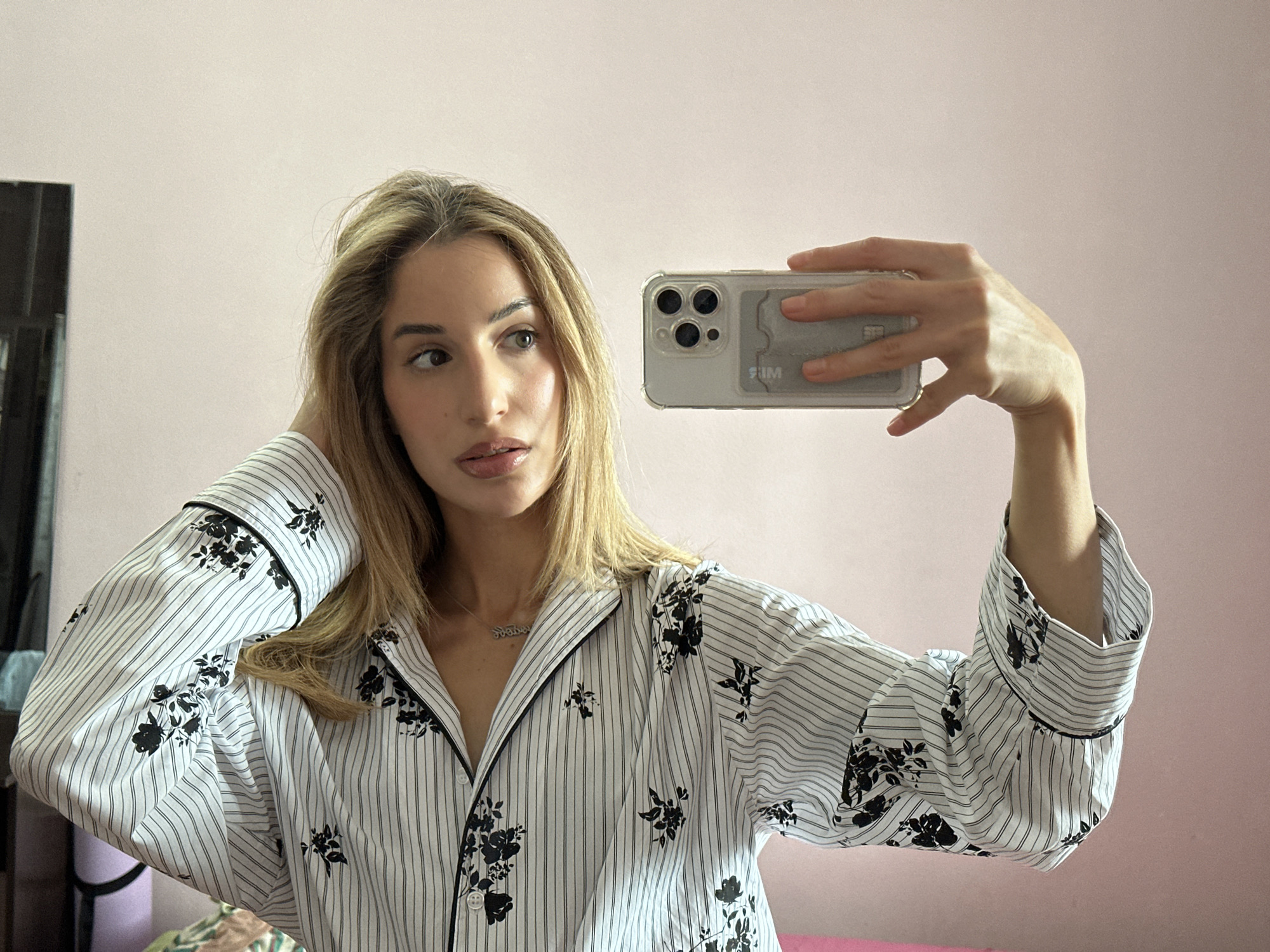 A woman with blonde hair takes a mirror selfie in a room with a pink wall. She is wearing a white pajama top with black pinstripes and black floral patterns. Her right hand is holding a smartphone, and her left hand is touching her hair. The room appears to be a bedroom, with a bed visible in the background. The image does not provide any information about the city of Moscow, Russia.