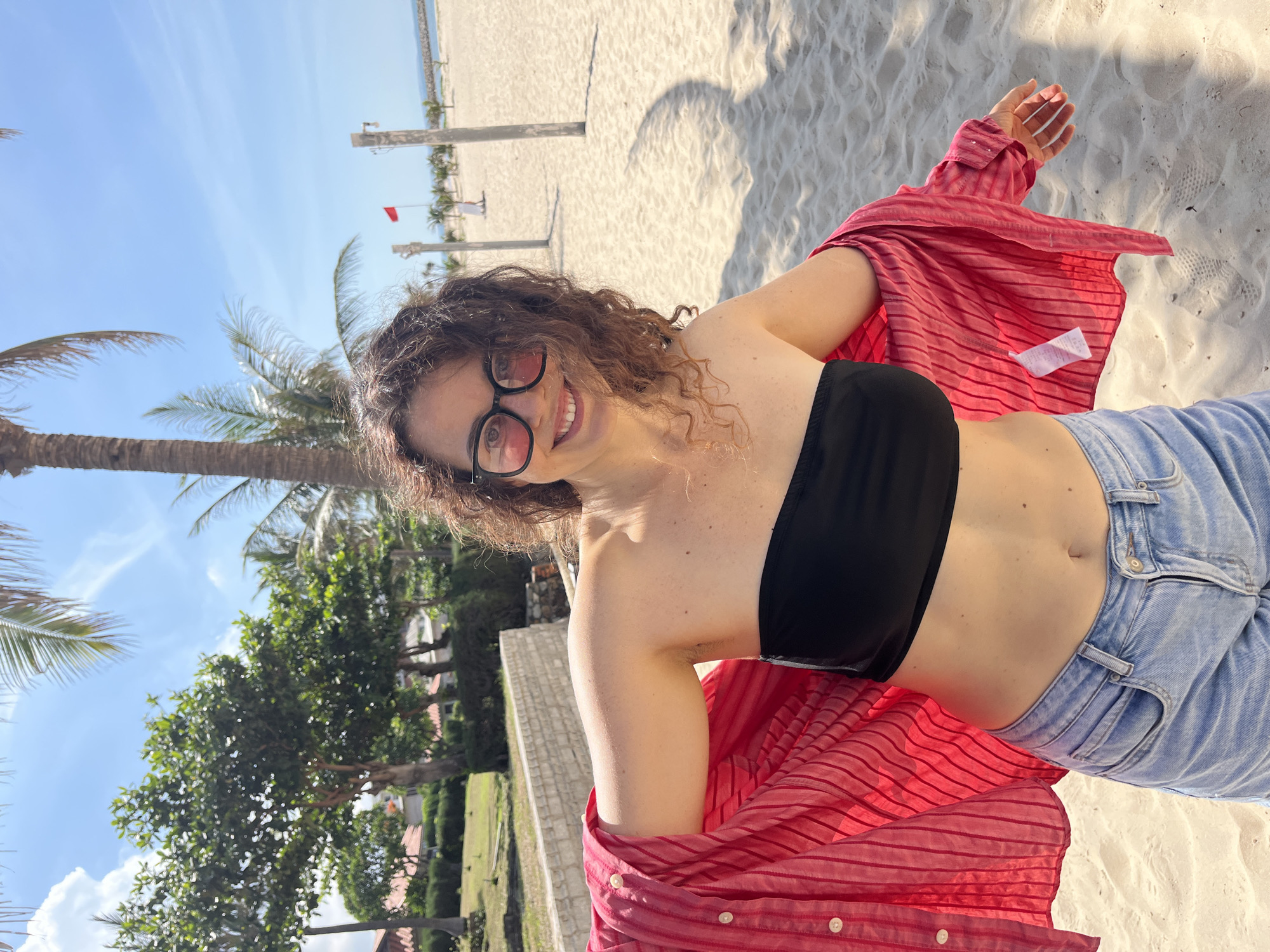 A woman stands on a sandy beach, her arms spread wide as she holds open a red, striped, unbuttoned shirt. She wears a black bandeau top and light blue denim shorts. She has curly brown hair and is wearing pink-tinted sunglasses. The background features a clear blue sky, palm trees, and a few buildings. The location is identified as Phu Thuy Ward, Vietnam.