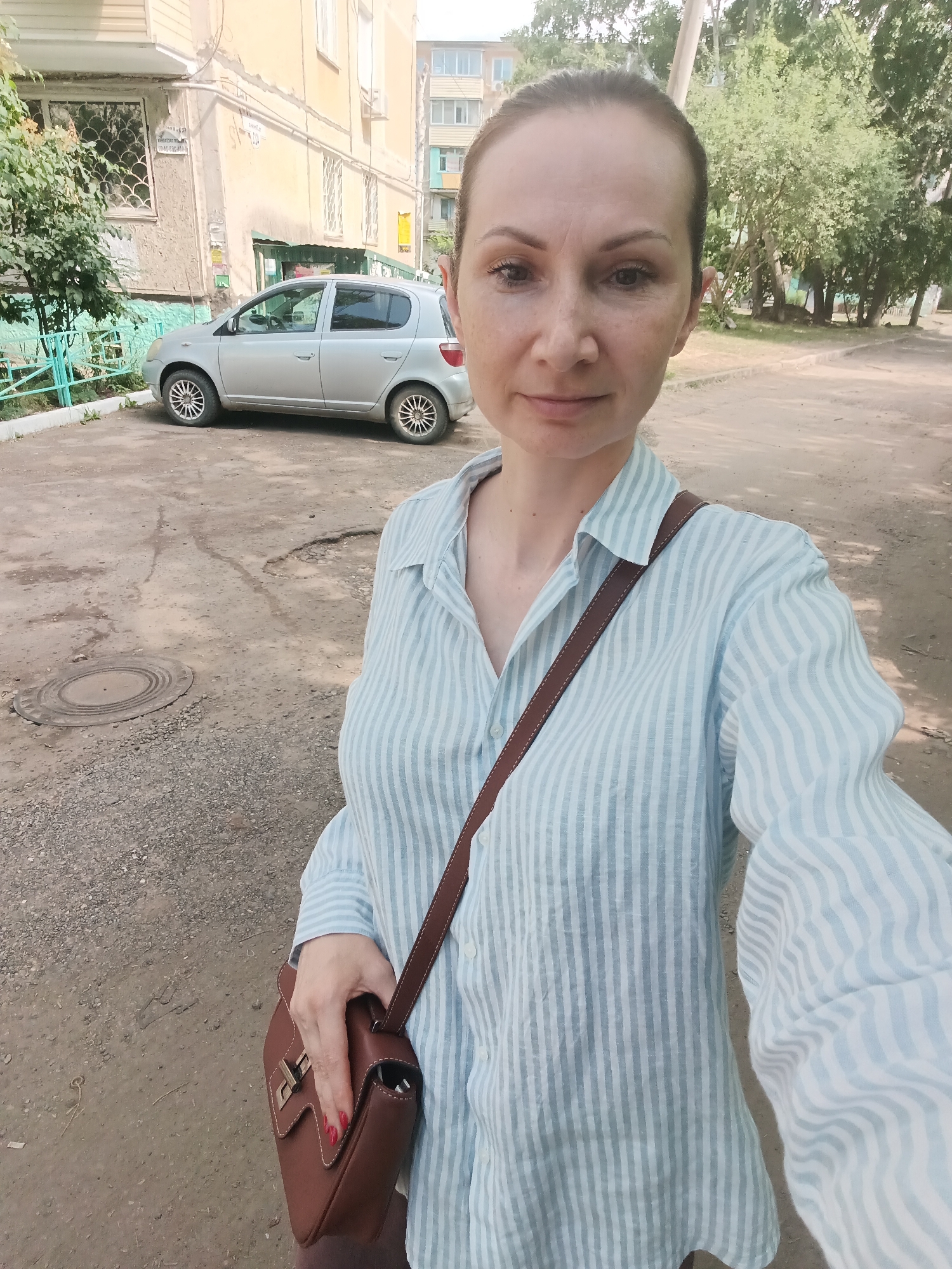 A woman takes a selfie outdoors in front of a pale yellow apartment building and a silver hatchback car. She is wearing a light blue and white striped button-up shirt and a brown leather crossbody bag. The ground is paved with asphalt and gravel, with some potholes visible. Trees and vegetation are present in the background. The location is identified as Komsomolsk-on-Amur, Russia.