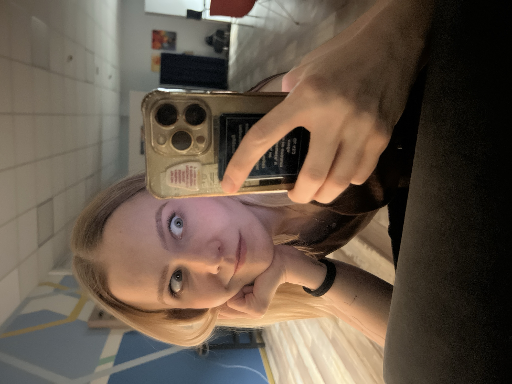 A woman with blonde hair and blue eyes is taking a selfie with a gold iPhone. She is holding the phone with her right hand, and her face is close to the camera. The background is a room with a bed, a wall with geometric patterns, and a hallway leading to another room with furniture. There is no indication of the location being Khimki, Russia.