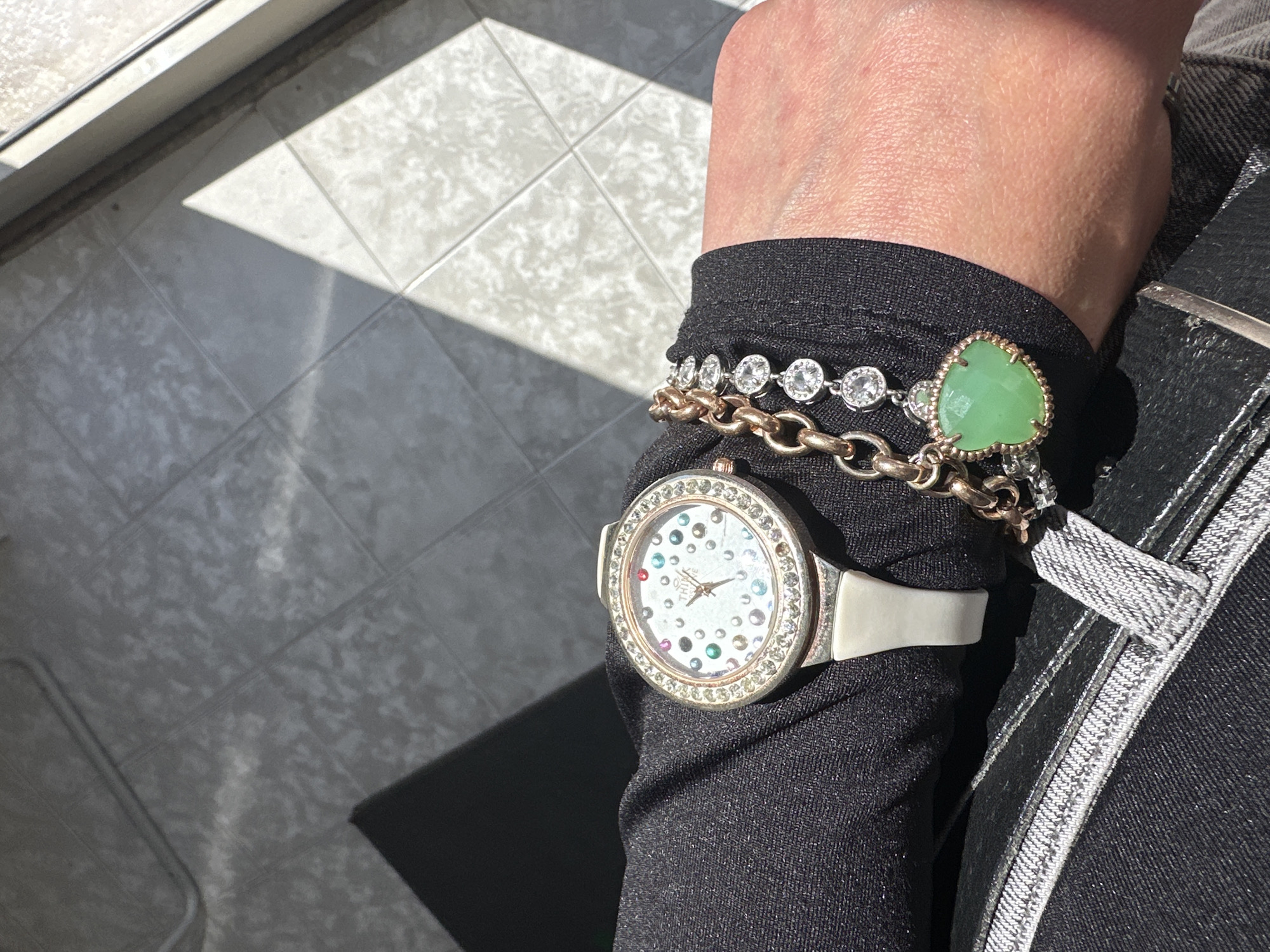 A close-up, high-angle shot captures a person's left wrist adorned with jewelry, set against a sunlit tiled floor in Venafro, Italy. The person wears a black long-sleeved top that covers part of their forearm, with their skin visible above the sleeve.
On the wrist, a white-strapped wristwatch features a circular, silver-toned bezel encrusted with clear rhinestones. Its white watch face is uniquely decorated with scattered small, multi-colored rhinestones in shades of red, green, blue, and clear. Gold-toned hands mark the time, and faint text, including "THE TIME," is visible on the face.
Above the watch, two bracelets are stacked. One is a sturdy gold-toned chain link bracelet. The second is a silver-toned bracelet with circular clear rhinestones, from which hangs a prominent heart-shaped pendant made of a light green stone, possibly jade or aventurine, framed by a silver or rose gold-toned border.
To the right, part of dark-colored (likely black) denim jeans is visible, characterized by light grey stitching or trim along a seam and pocket area.
The background shows a light grey or off-white tiled floor with a mottled texture. Bright sunlight streams in from the upper left, casting sharp, geometric shadows across the floor, indicative of daytime. A dark, rectangular mat or rug is partially visible on the lower left of the floor. A metallic or uPVC frame of a window or door is evident along the upper left edge, marking the source of the strong natural light.