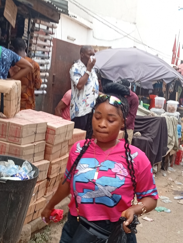 A young woman in a pink shirt with birds and the number 23 walks through a bustling outdoor market. She has braided hair and sunglasses on her head. Behind her, a man in a patterned shirt holds a tissue to his face. Boxes are stacked to the left of the woman near a black trash can, and market stalls with umbrellas are visible in the background. The ground appears littered with trash.